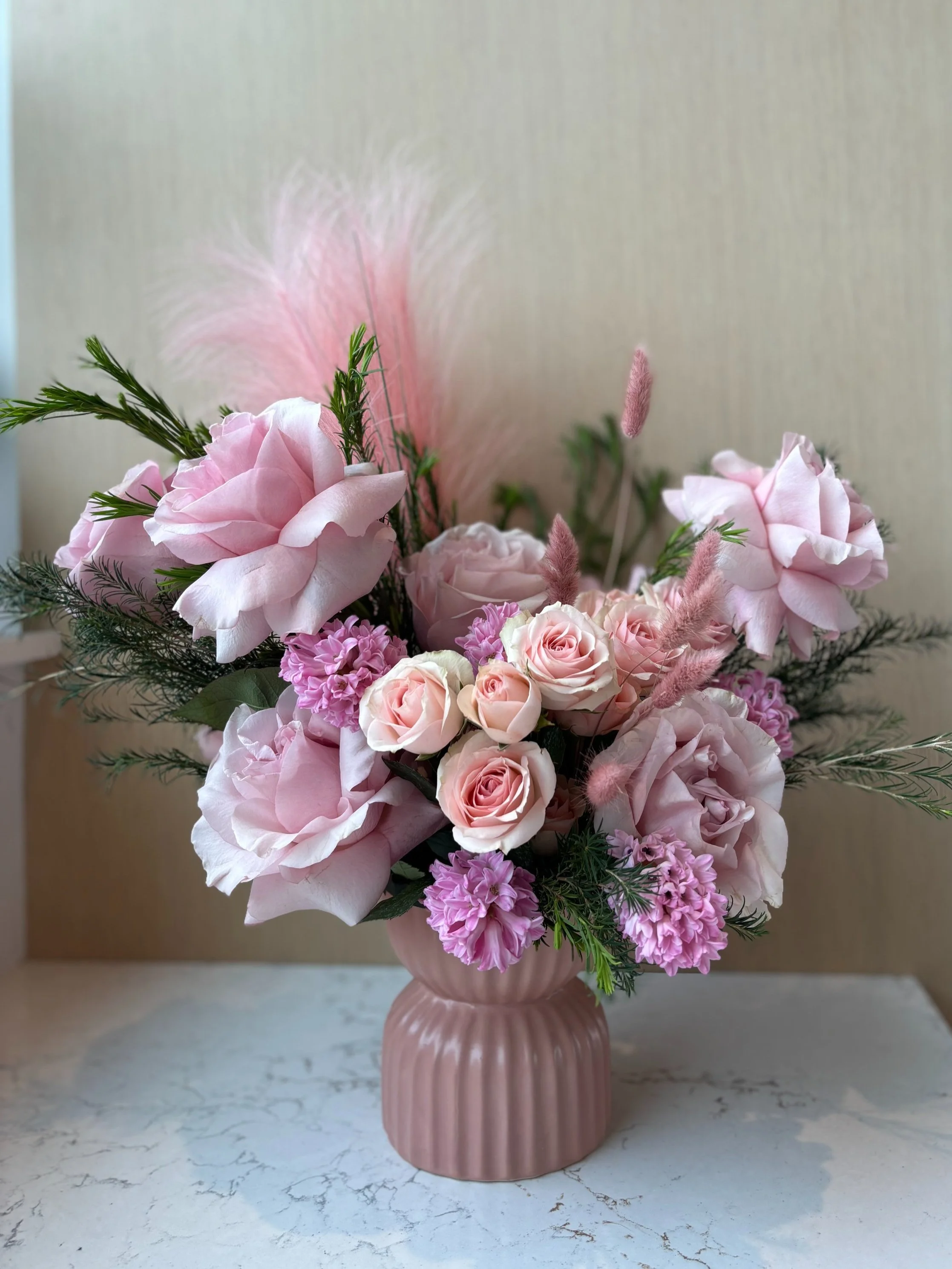Pink floral bouquet with roses, peonies, lavender, and greenery in a pink vase on a white marble surface.