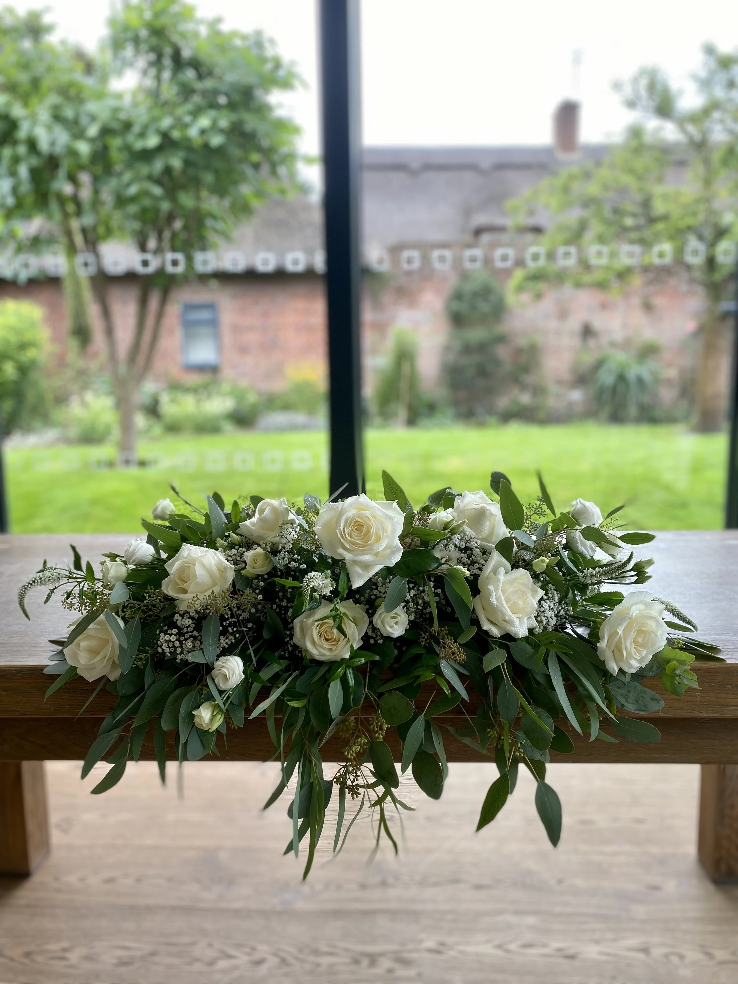 A white floral arrangement with roses and greenery on a wooden table in front of a window showing a garden.