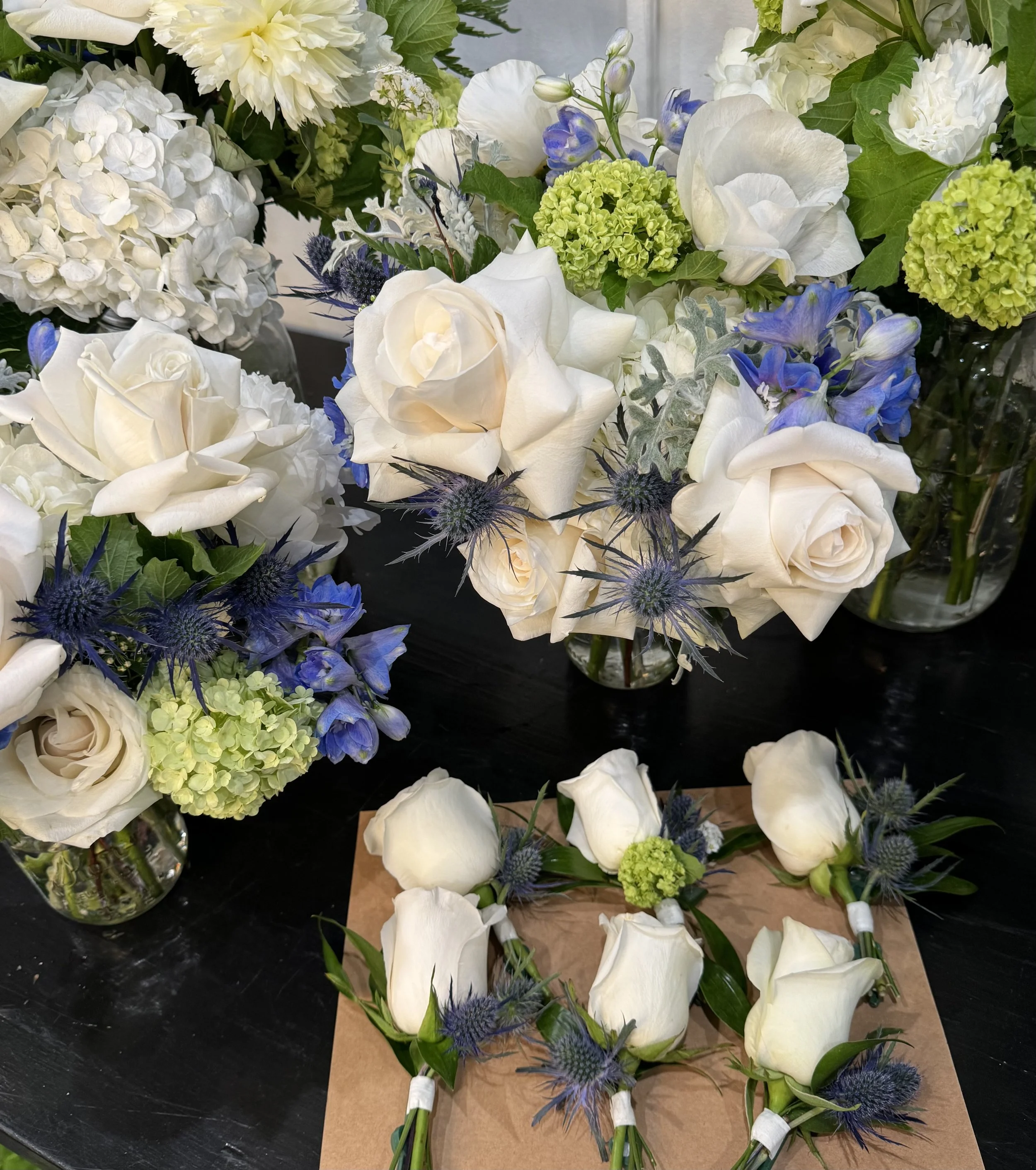 Various bouquets of white roses, hydrangeas, blue thistles, and green hydrangeas arranged in vases and on a table, with some boutonnières assembled with white roses, blue thistles, and greenery.