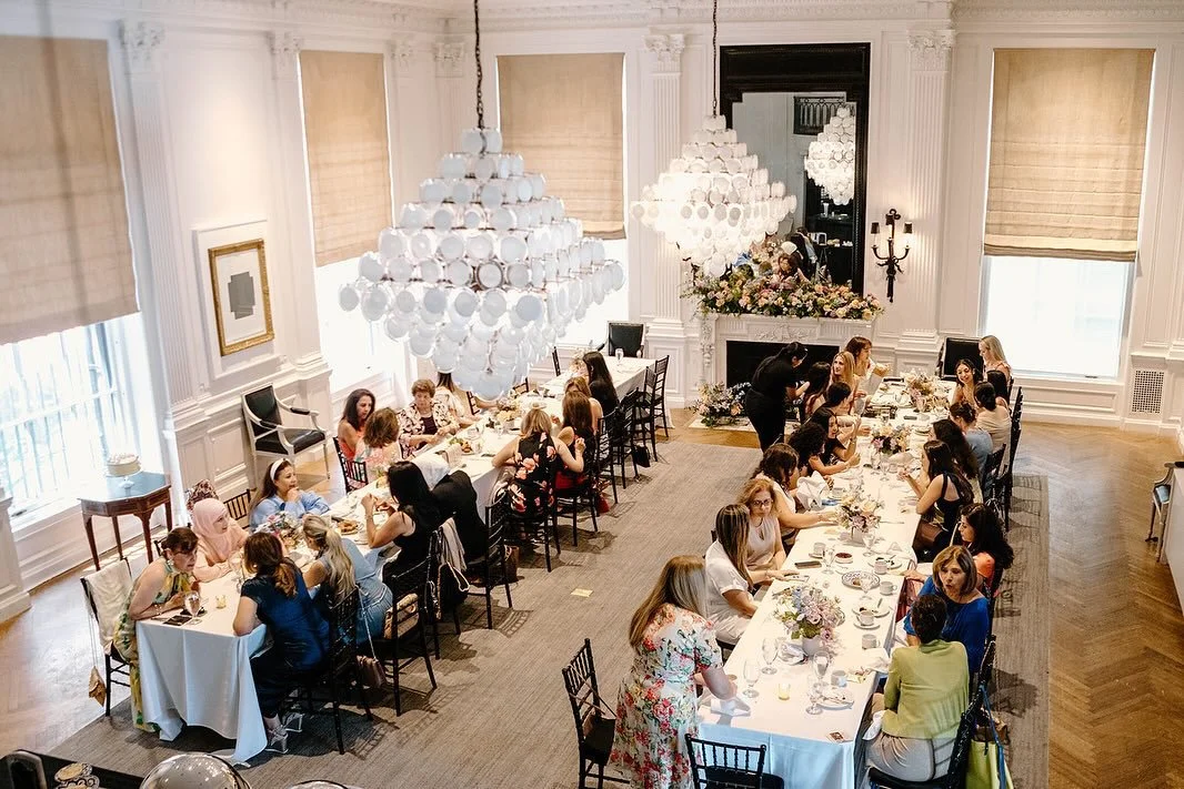 A large, elegant dining room with white walls, ceiling moldings, and tall windows with beige shades. Two large chandeliers hang above a long banquet-style table, which is decorated with floral centerpieces. Many women are seated around the table, soc