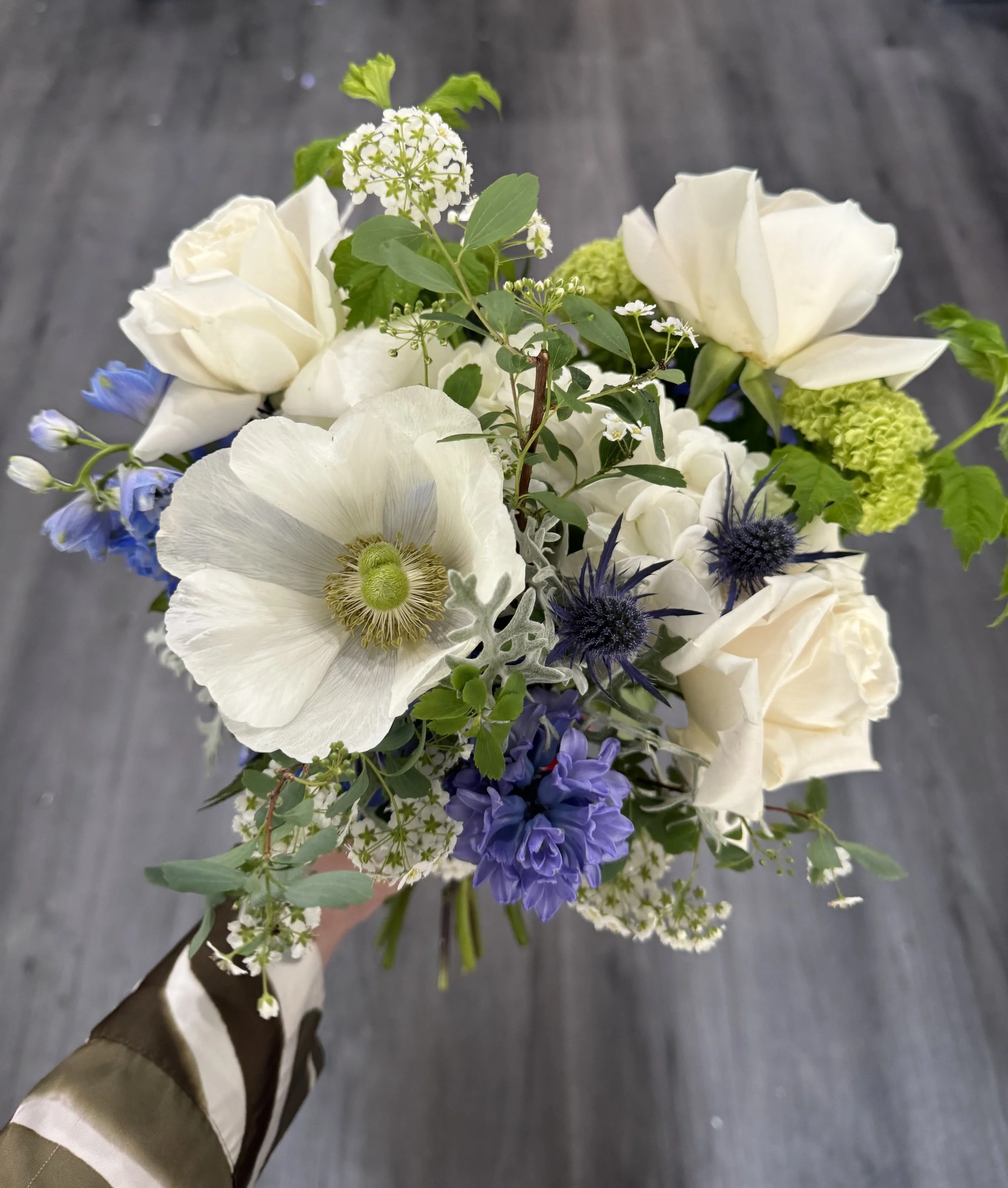 Hand holding a bouquet of white, purple, and green flowers against a gray wooden floor background.