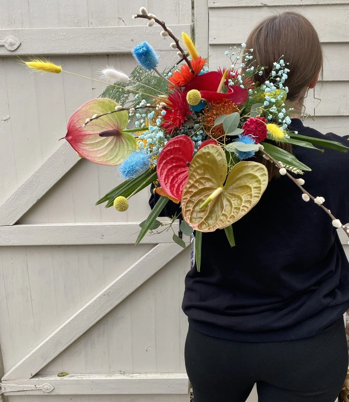 Person holding a large, colorful floral bouquet with various exotic flowers and greenery against a white wooden fence background.