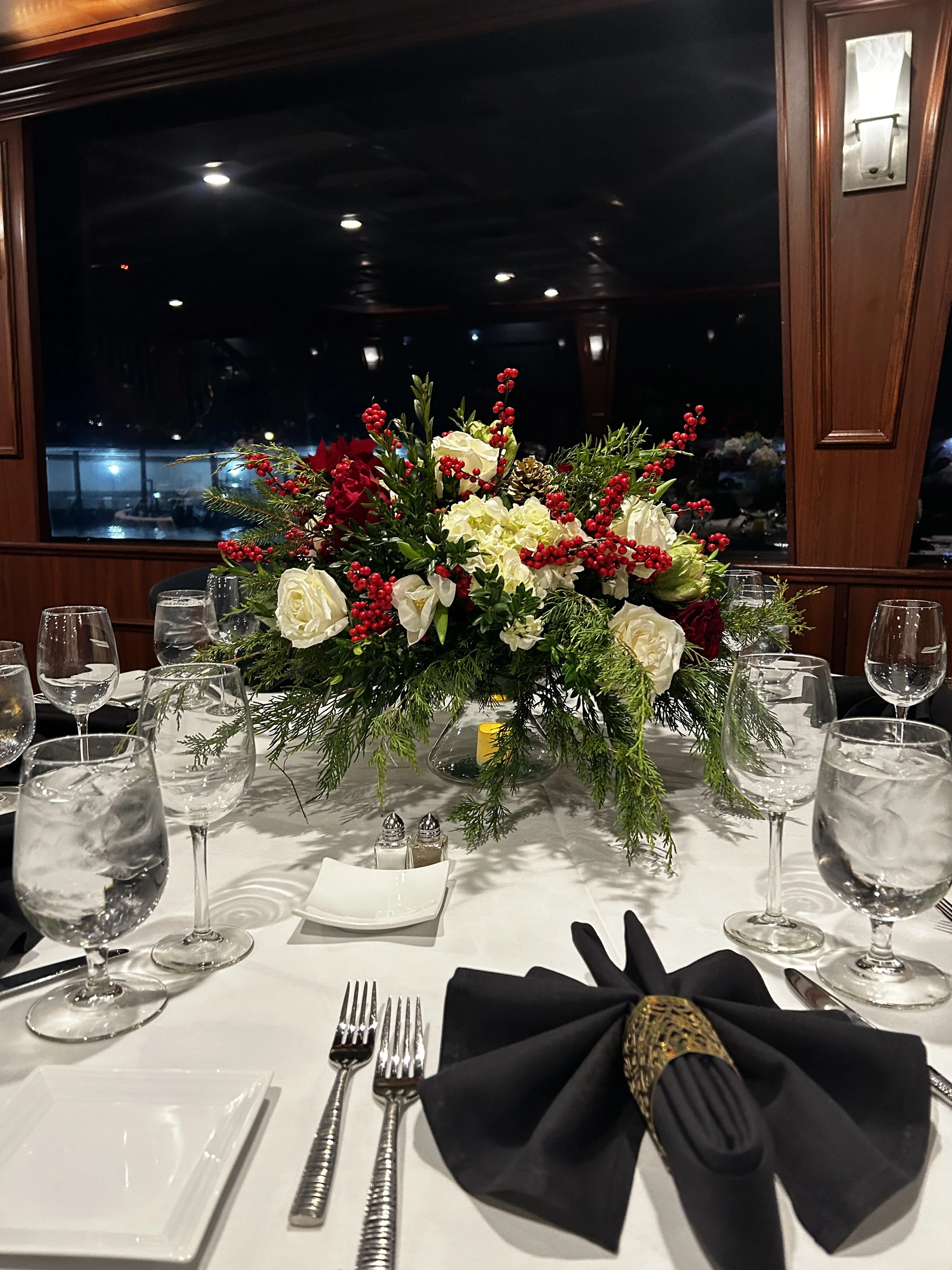 Elegant dining table with a large floral centerpiece consisting of white roses, red berries, and greenery, set with glasses, plates, and black napkins with gold napkin rings.