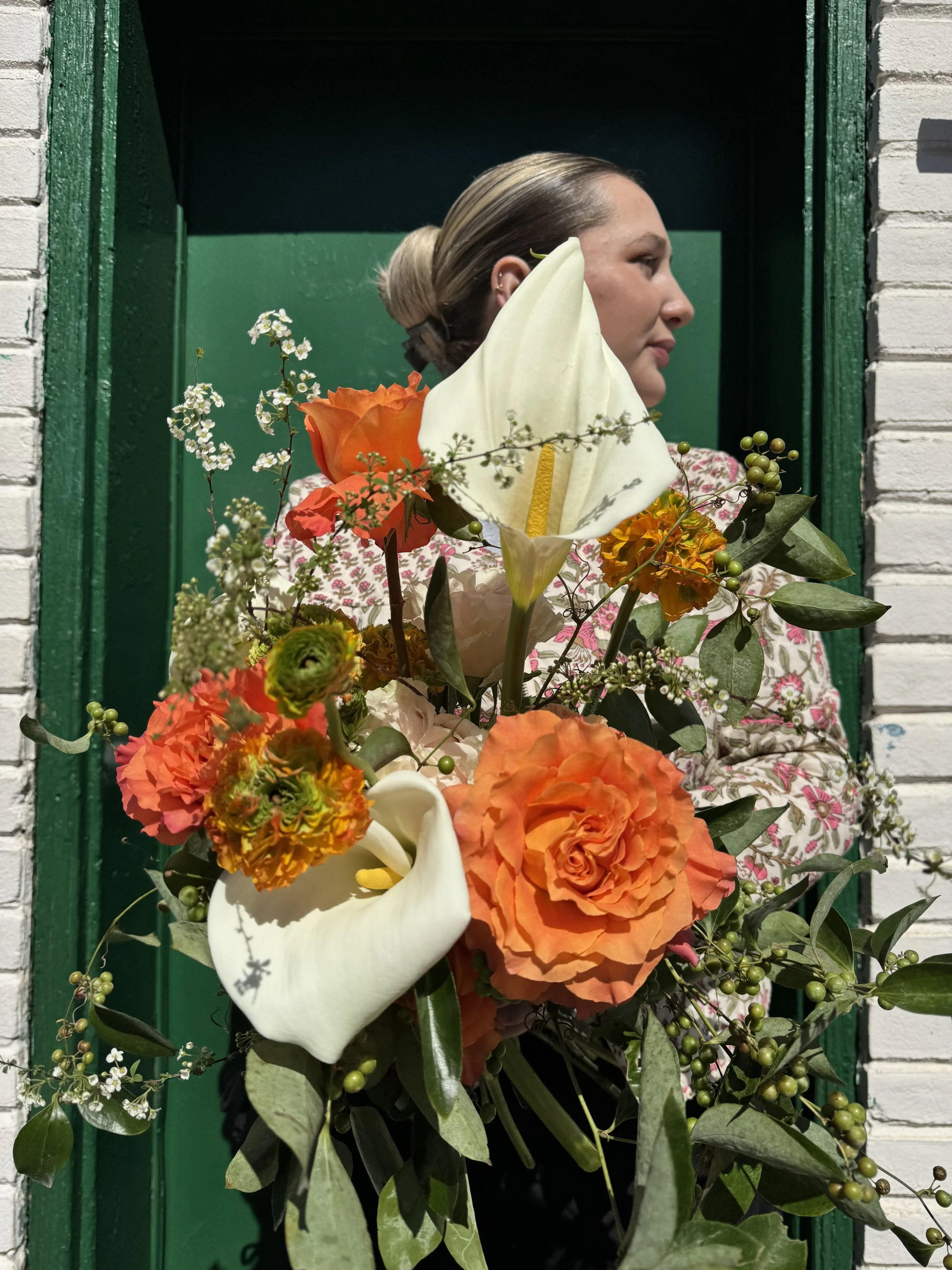 A woman holds a large bouquet of colorful flowers, including white calla lilies, orange roses, and smaller orange and white blossoms, in front of a green door and a white brick wall.