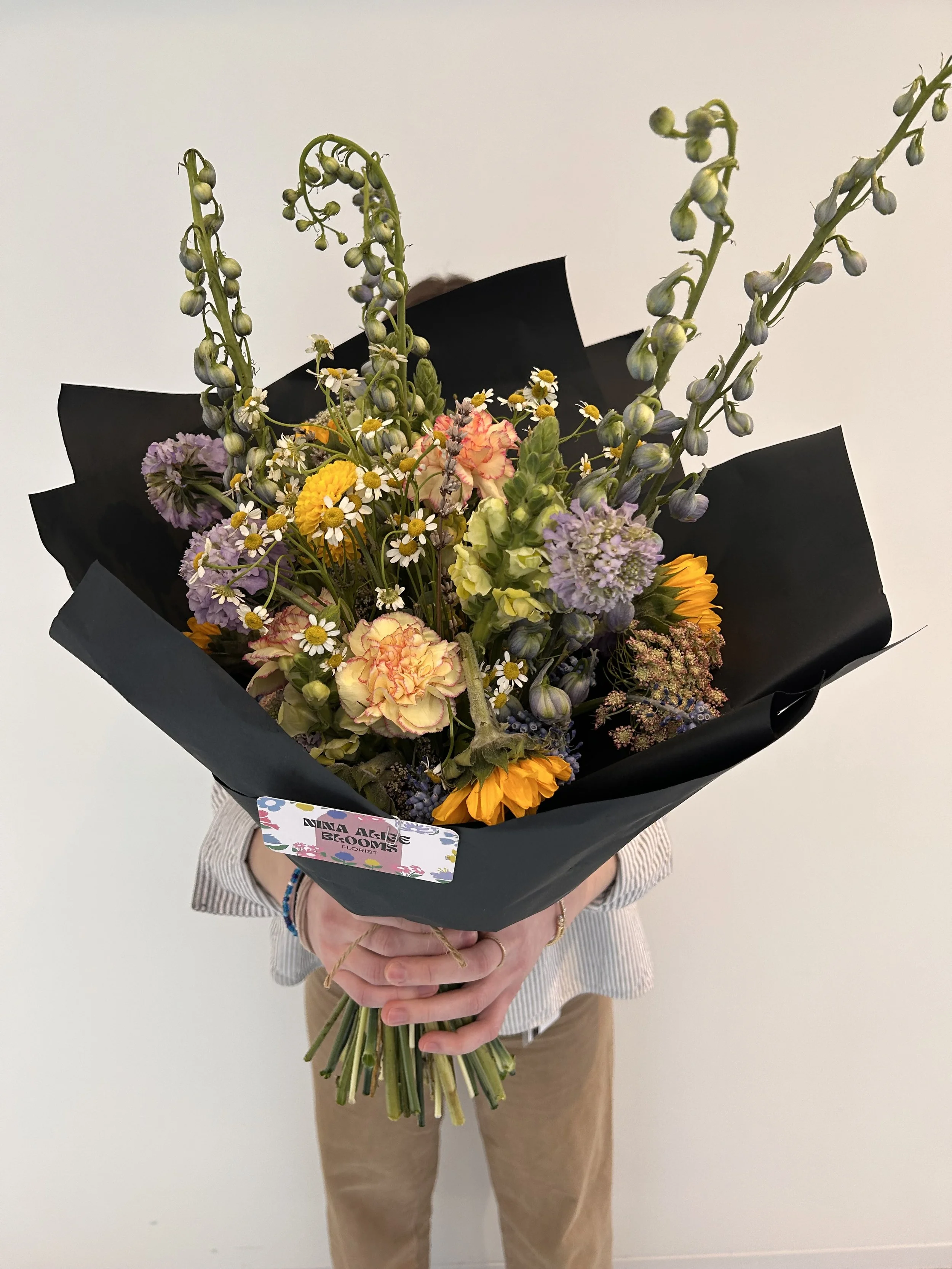 Person holding a bouquet of colorful flowers wrapped in black paper against a plain white background.