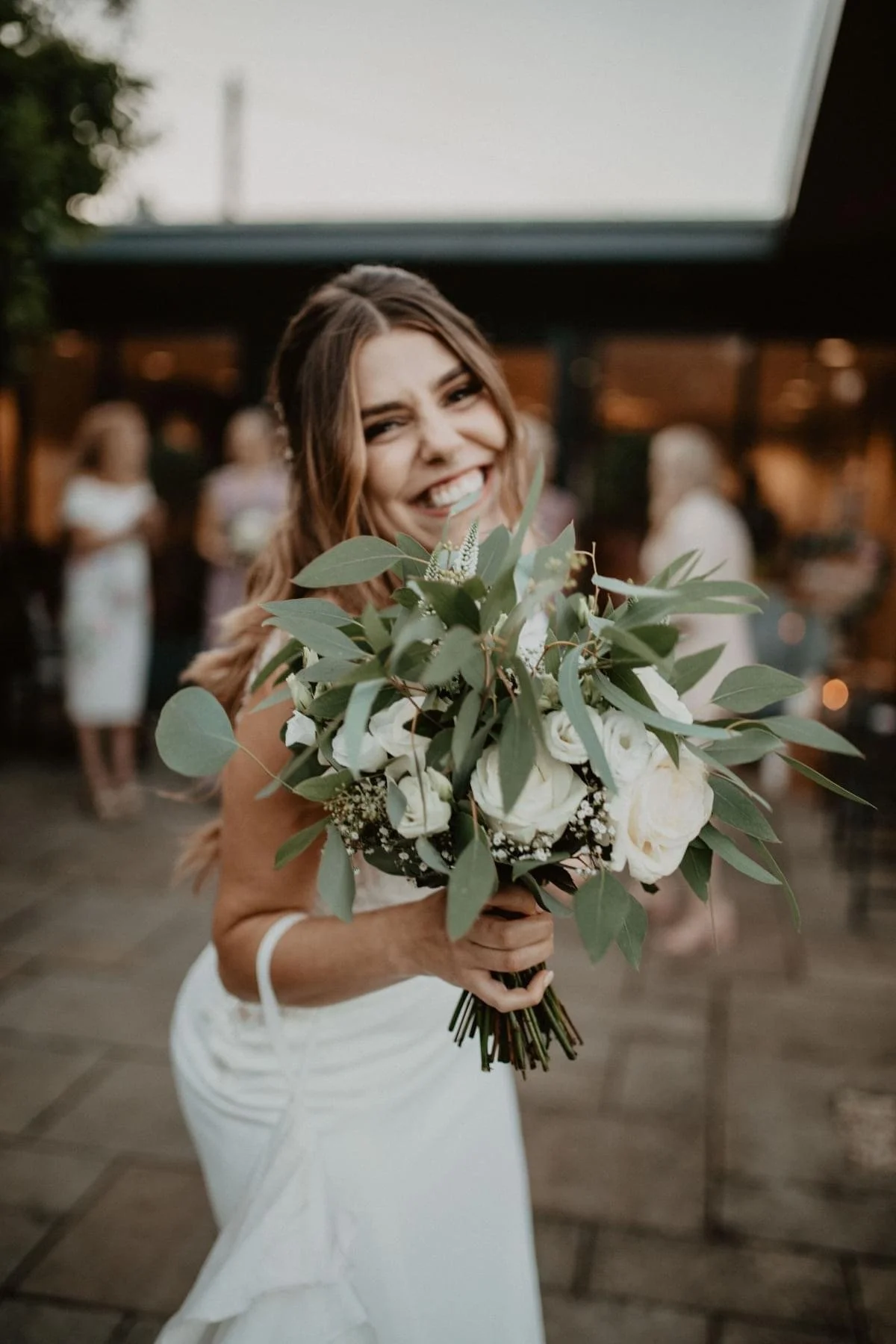 Bride in a white dress smiling and holding a bouquet of white roses and green leaves at a wedding reception.