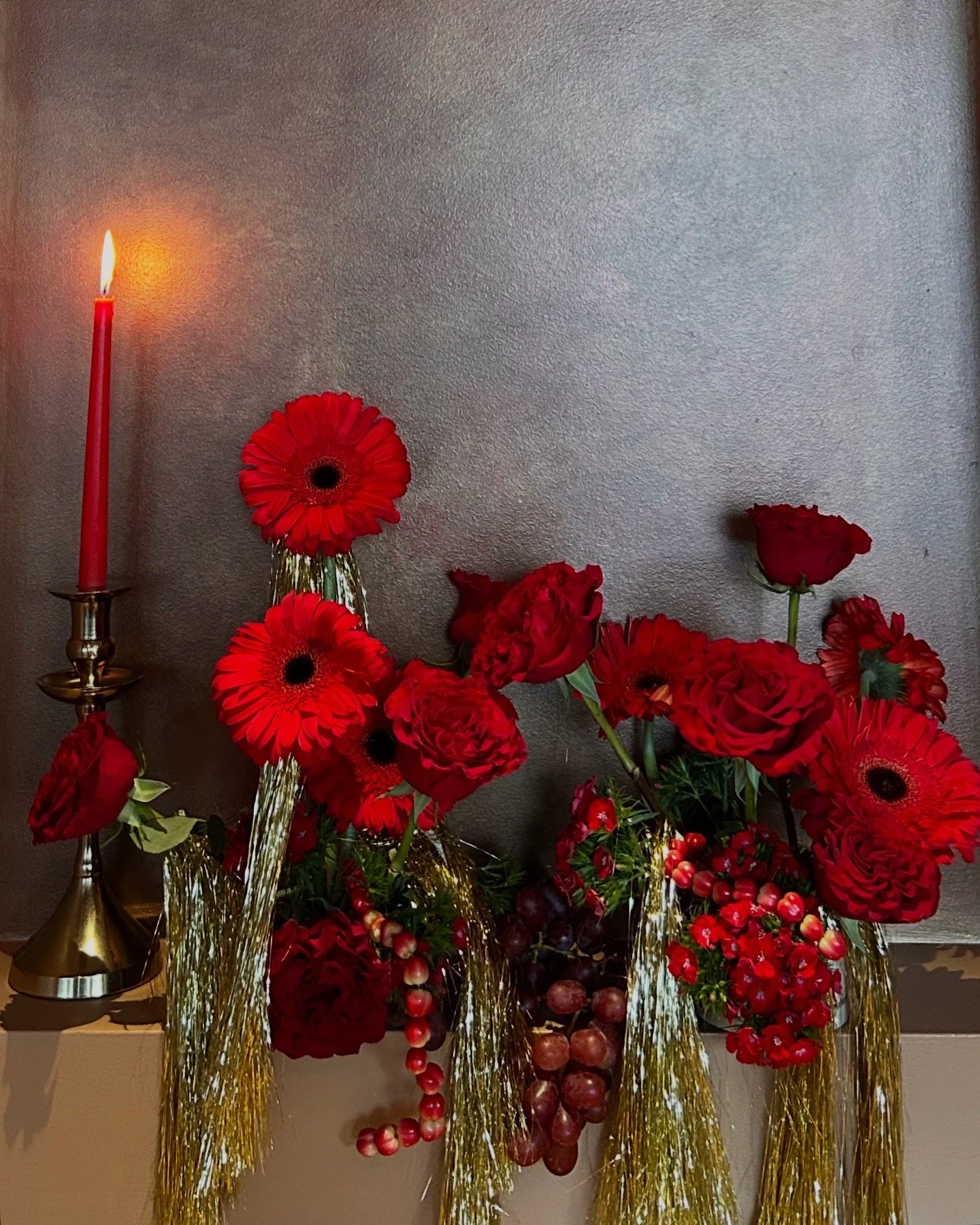 Red flower arrangement with roses and gerbera daisies in gold vases, a red candle on a gold candlestick, and strings of grapes and decorative gold strands against a gray wall.