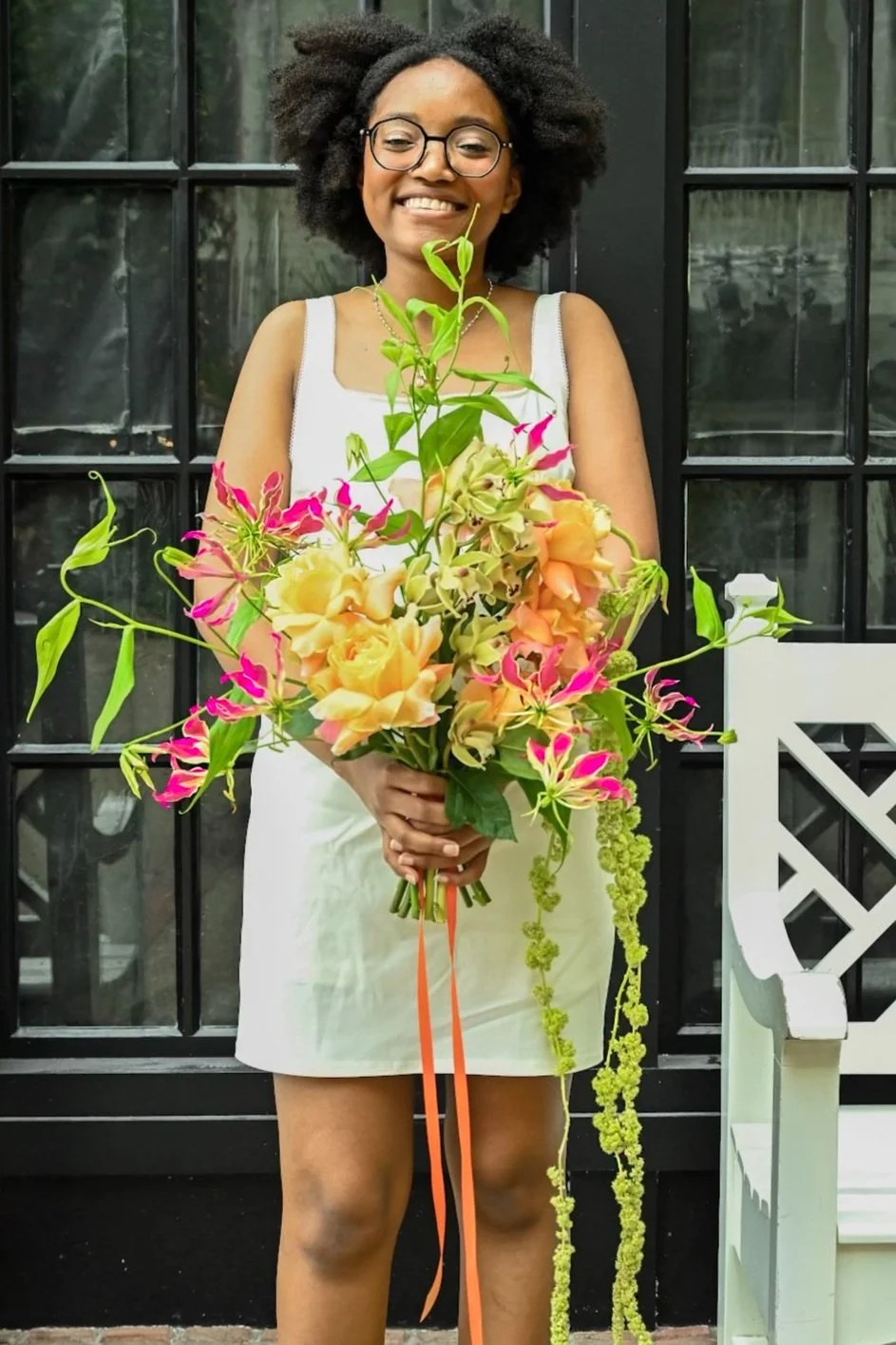 A woman with glasses and curly hair smiling and holding a large bouquet of colorful flowers, standing in front of a black windowed door.