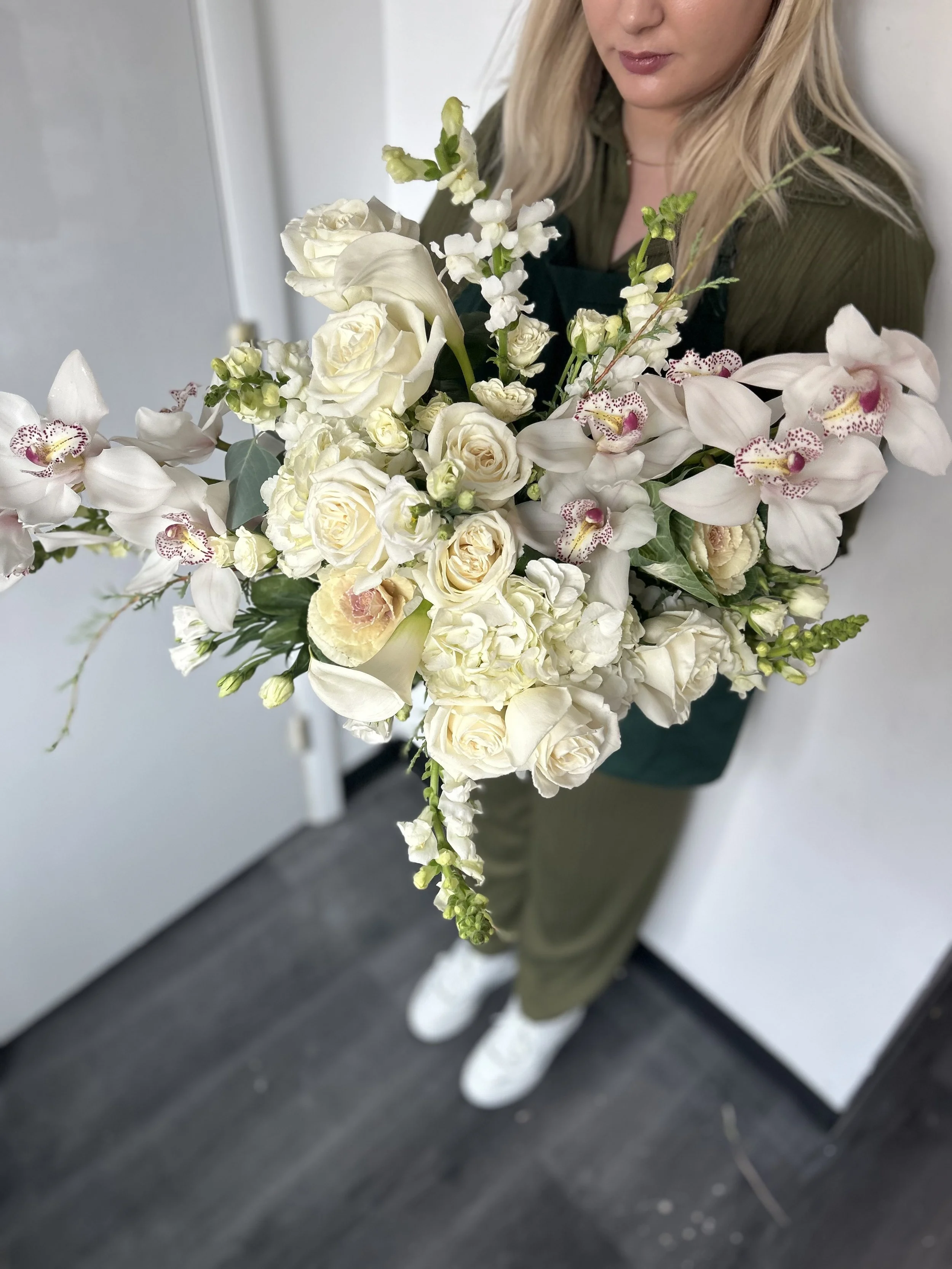 A woman holding a large bouquet of white and cream-colored flowers including roses, orchids, and other flowers, with a white wall and door in the background.