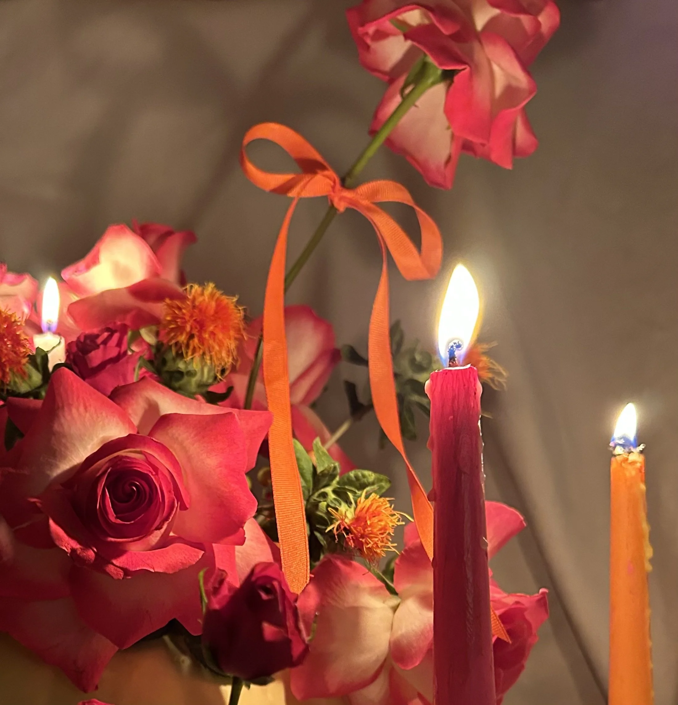 A bouquet of pink and red roses with orange ribbons, lit with two tall pink and orange candles.