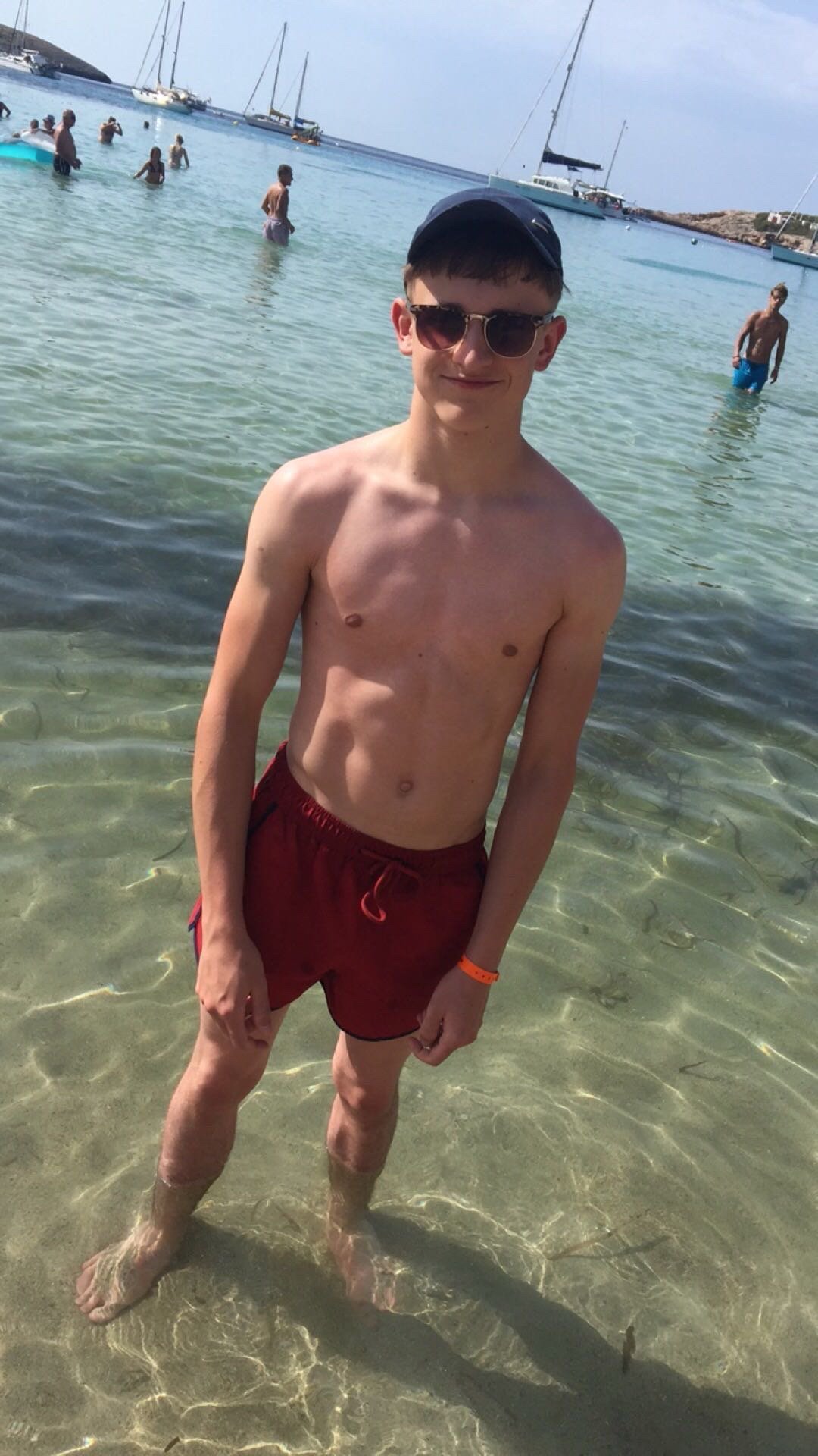 A young man with sunglasses, a black cap, red shorts, and an orange wristband standing in shallow clear water at a beach with sailboats in the background.