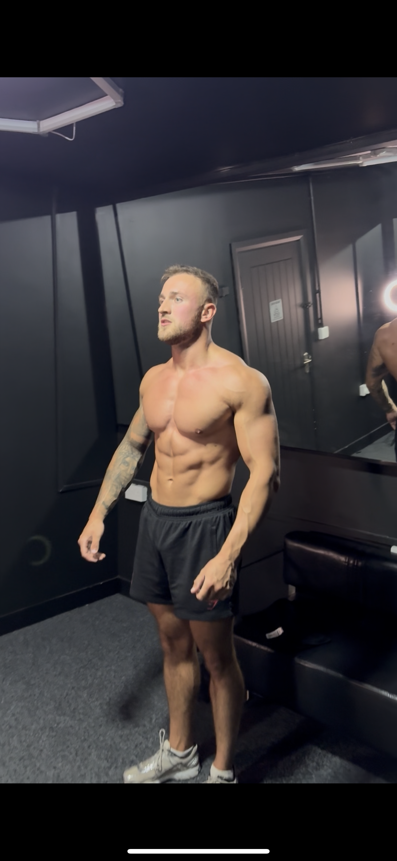 A shirtless man with athletic build, tattooed arms, wearing black shorts and white sneakers, standing in a room with black walls and a mirror.