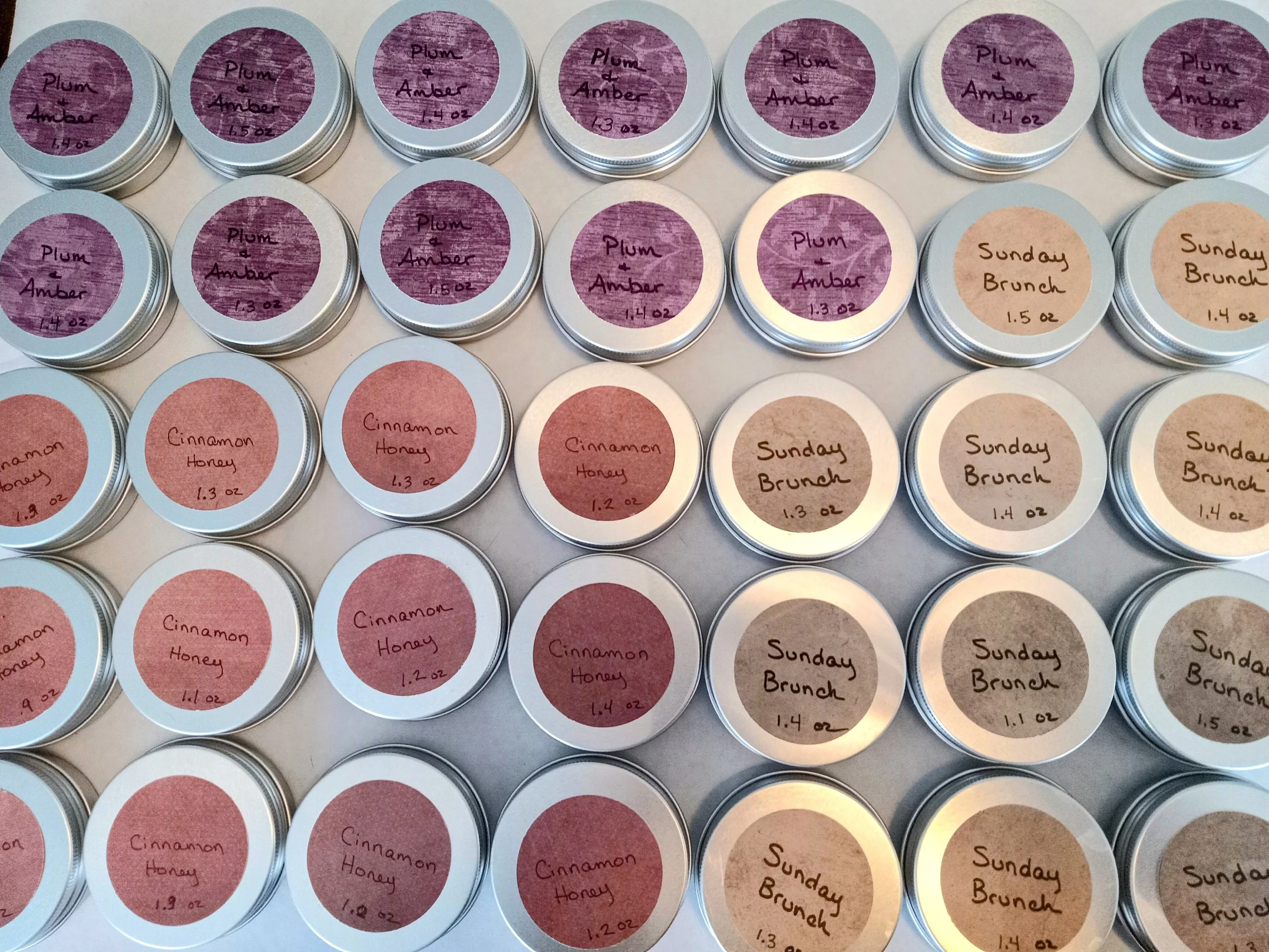 Image shows five rows of round silver tins containing lotion bars on a white background. The labels say Plum and Amber, Sunday Brunch, and Cinnamon Honey.