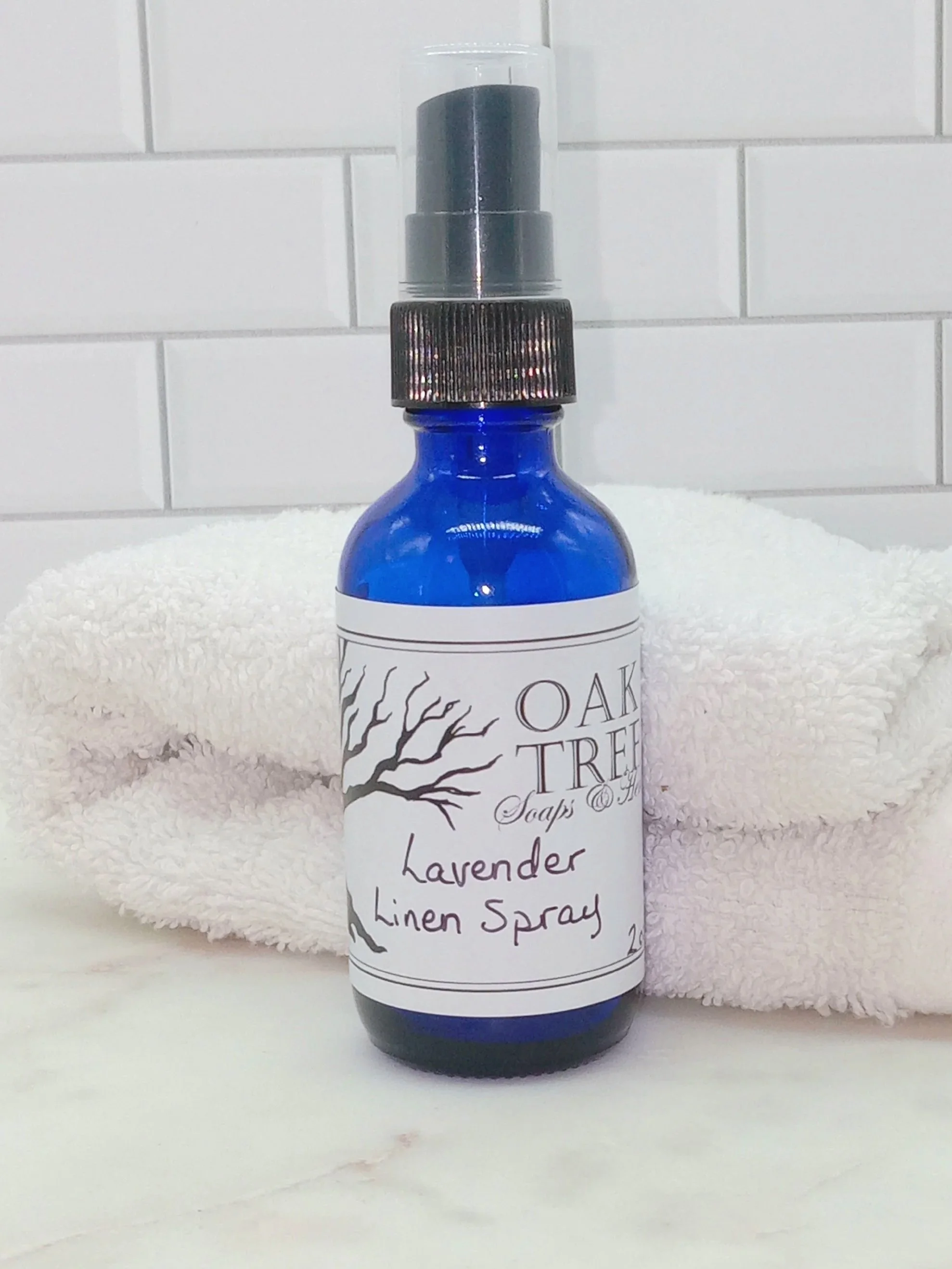Image shows a close up picture of a dark blue glass bottle with a black spray top.  The label says Lavender Linen Spray.  The bottle is in front of a folded white towel and white tile background.