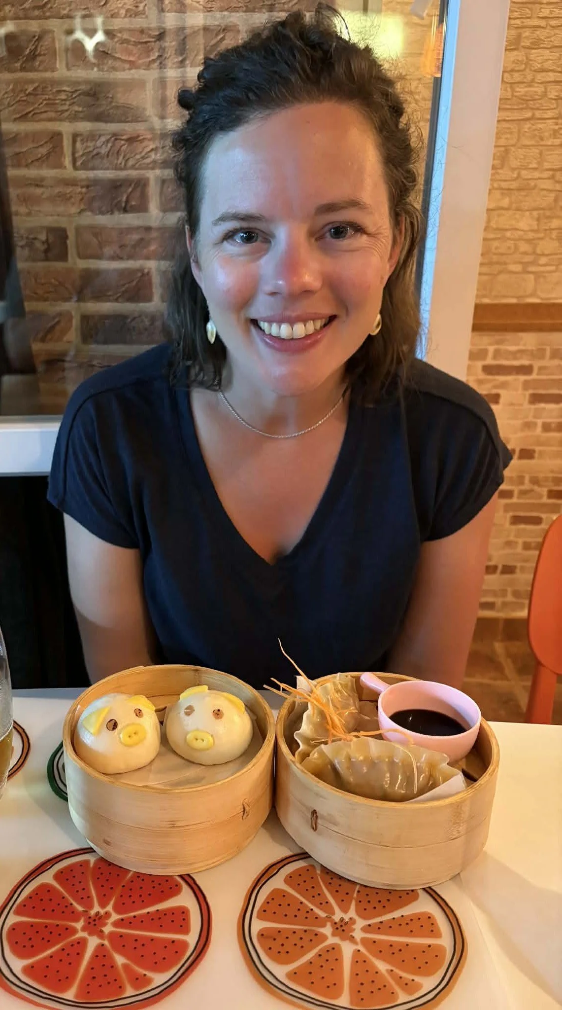 A woman sitting at a table with two steamer baskets of food, smiling at the camera. One basket contains steamed buns decorated to look like pigs, and the other contains dumplings with a dipping sauce. The table has orange and black patterned placemats resembling grapefruit slices.