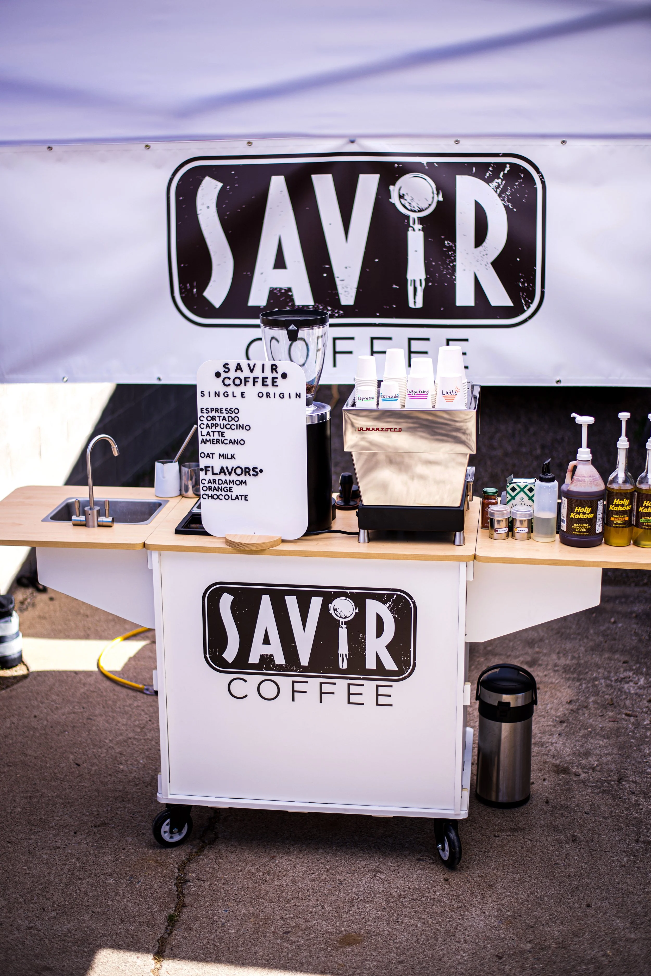 A mobile coffee stand with a black and white sign that reads 'SAVIR COFFEE' above the setup. The stand features a menu listing single origin coffee options and flavors, a small sink, disposable coffee cups, syrup bottles, and a coffee grinder.