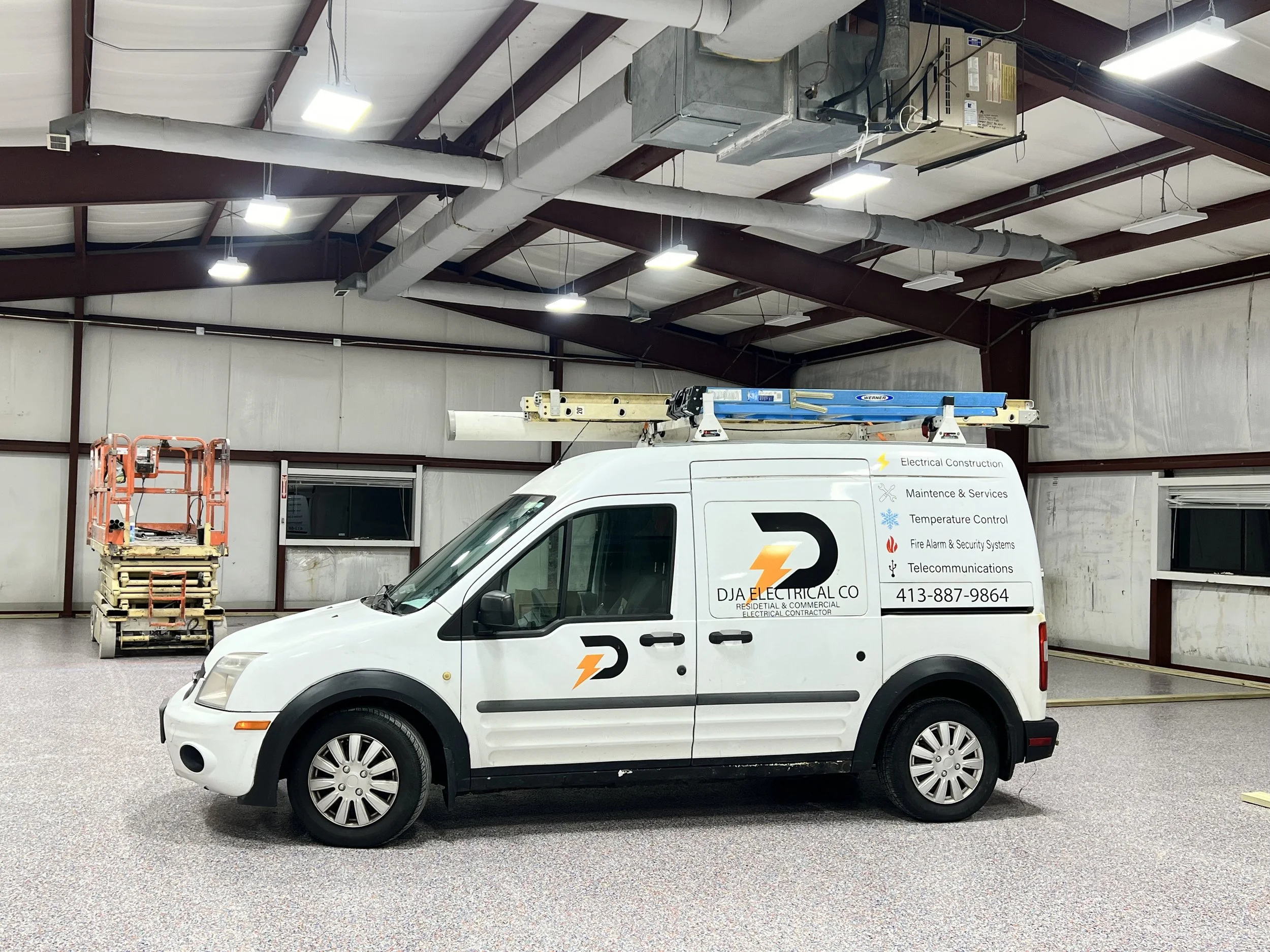 White electrical service van inside a large industrial warehouse with a scissor lift in the background, displaying company's services including electrical construction, maintenance, temperature control, fire alarm, security systems, and telecommunications.