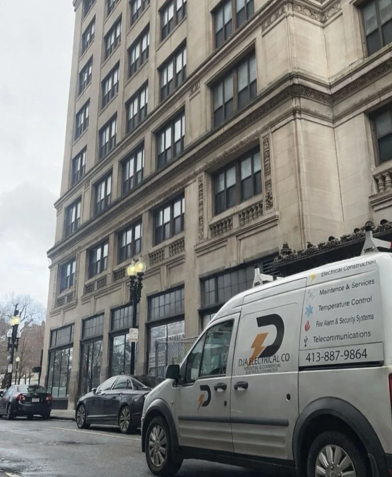 A city street with parked cars in front of a large multi-story building with ornate architectural details. A service van labeled for electrical, security, and telecommunications work is parked in the foreground.