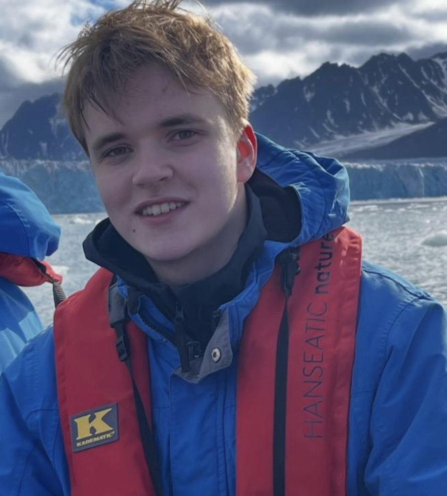 Young man with light brown hair smiling, wearing a blue jacket and red life vest, with snowy mountains and glaciers in the background.