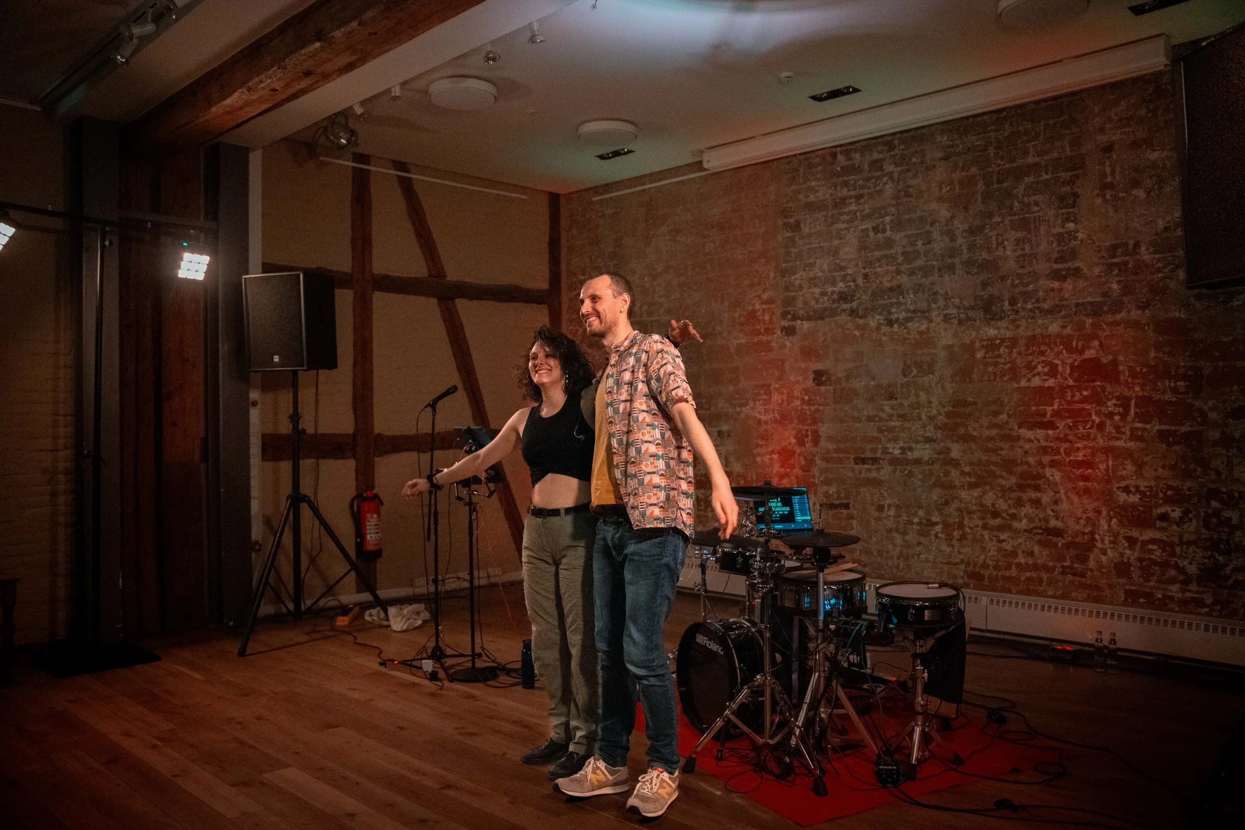 Zwei Menschen stehen auf einer Bühne in einem Raum mit Backsteinwand, sie sind lächelnd und scheinen sich zu amüsieren, es gibt Bühnenmusik-Equipment im Hintergrund.