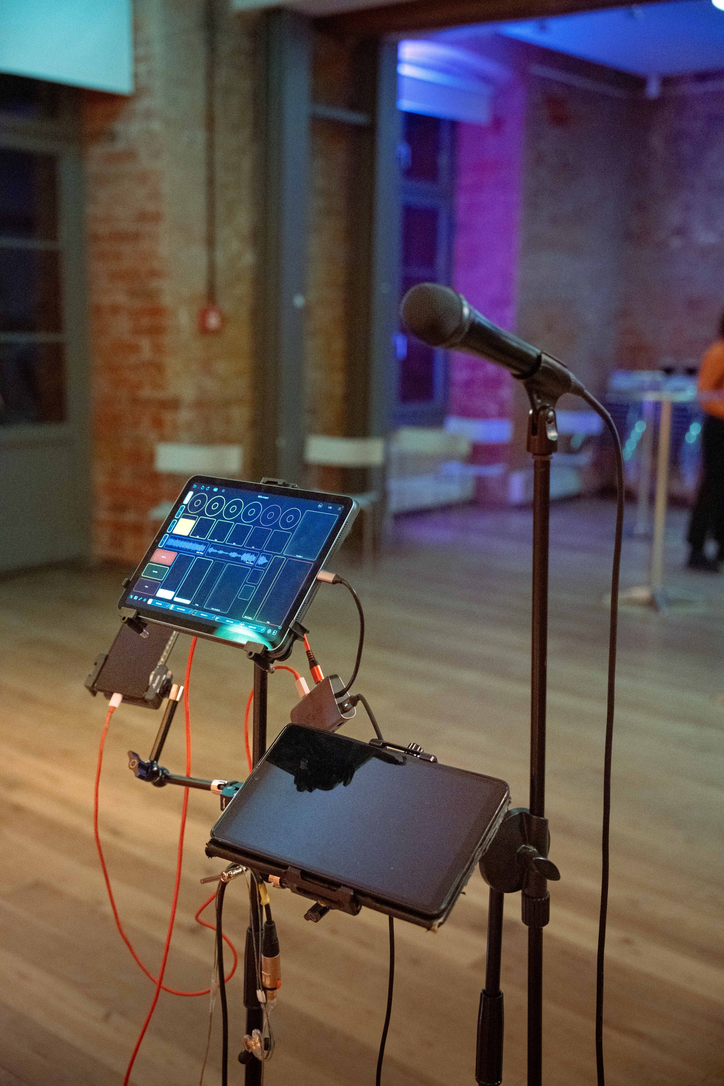 Ein Mikrofonständer mit einem Mikrofon und einem Tablet, das Musiksoftware zeigt, in einem Raum mit Holzfußboden und einer Backsteinwand im Hintergrund.