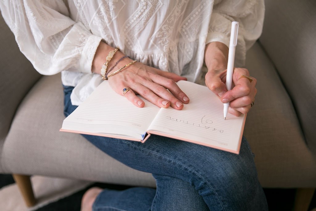 Person sitting on a couch, writing in a notebook with a white pen. They are wearing a white blouse with lace details and jeans, with several bracelets and rings on their hands.