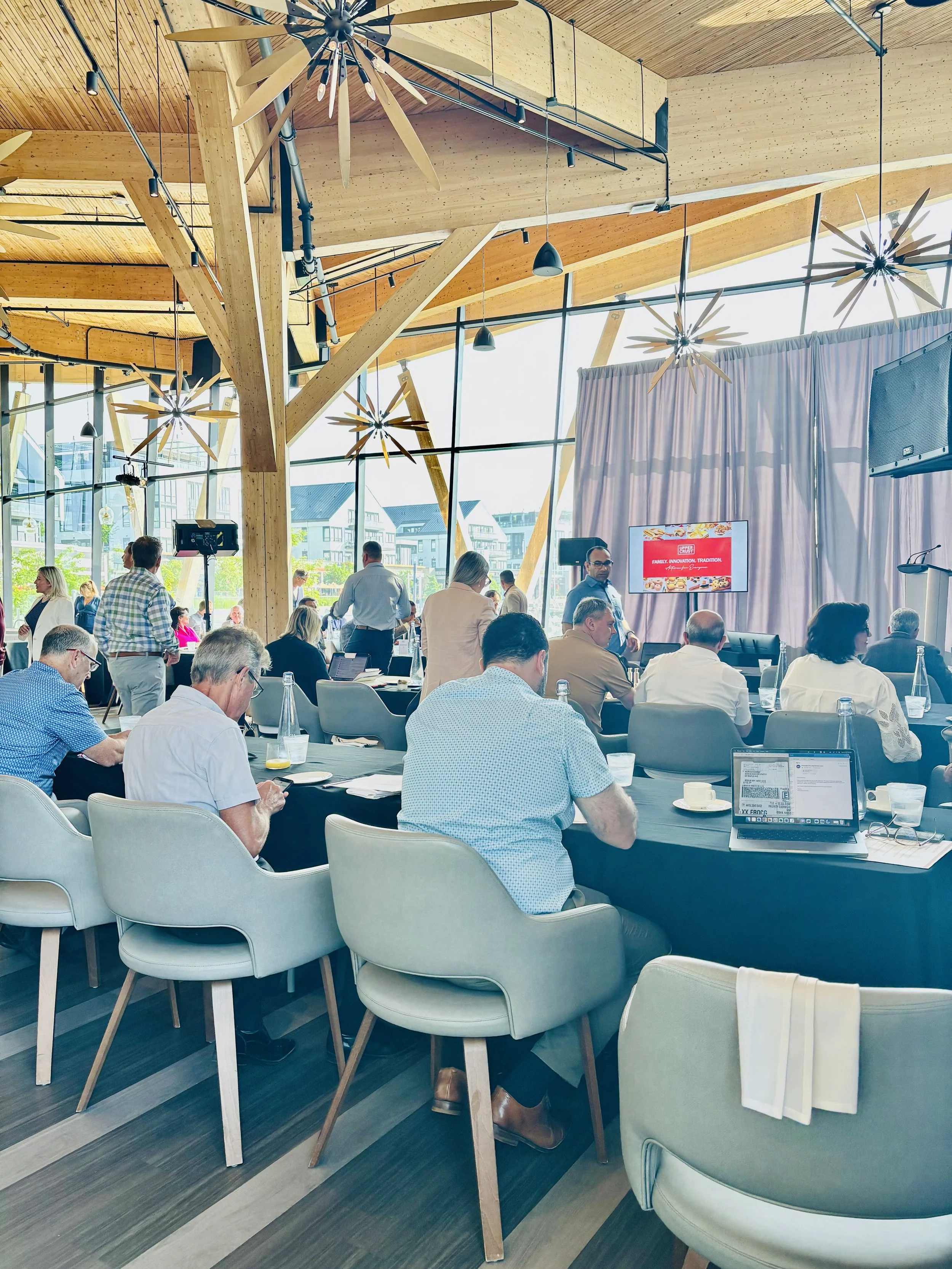 A conference room filled with people sitting at tables, some working on laptops, with large windows letting in natural light. The ceiling has wooden beams and decorative lighting fixtures.