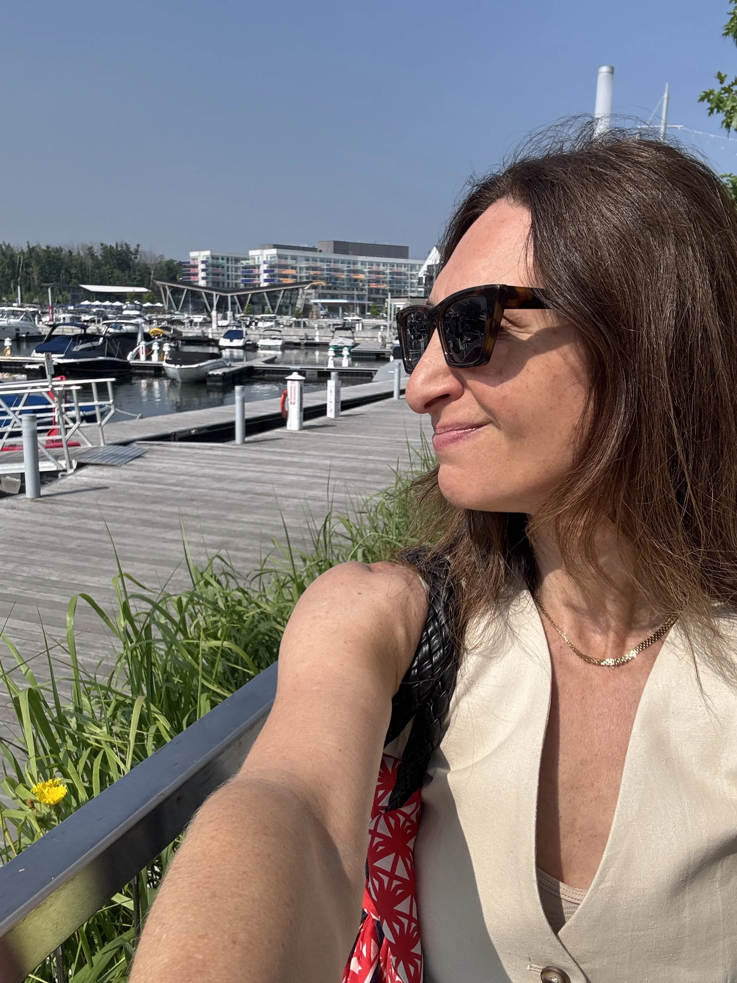 A woman wearing sunglasses and a light-colored top taking a selfie at a marina with boats docked and modern buildings in the background on a sunny day.