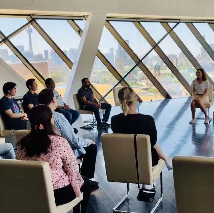 A diverse group of people attending a small indoor seminar or workshop in a modern room with large windows showing a city view.