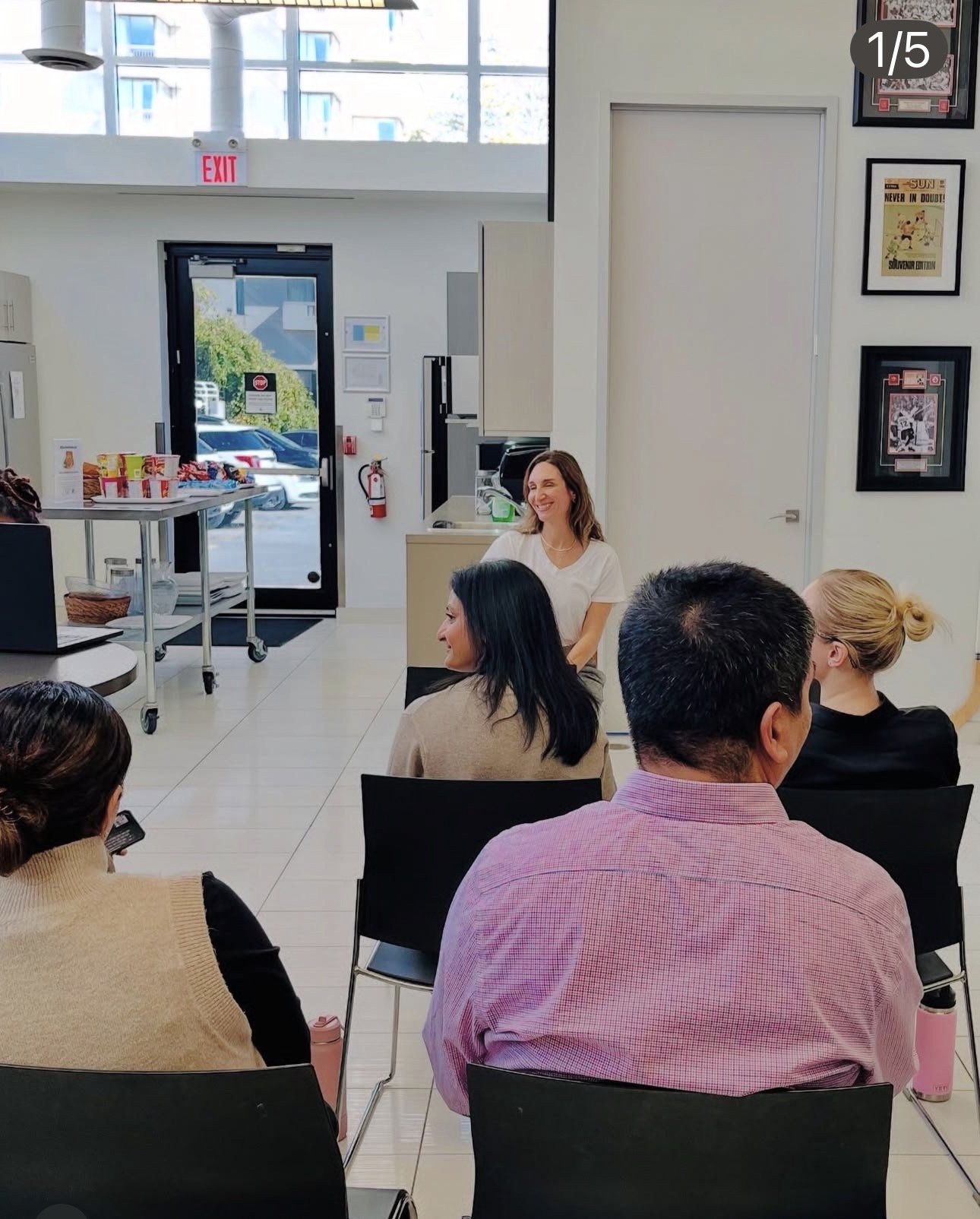 A group of people sitting in chairs in a room with white walls and artwork, listening to a woman who is standing and smiling, in an office or seminar setting with large windows and an exit door in the background.