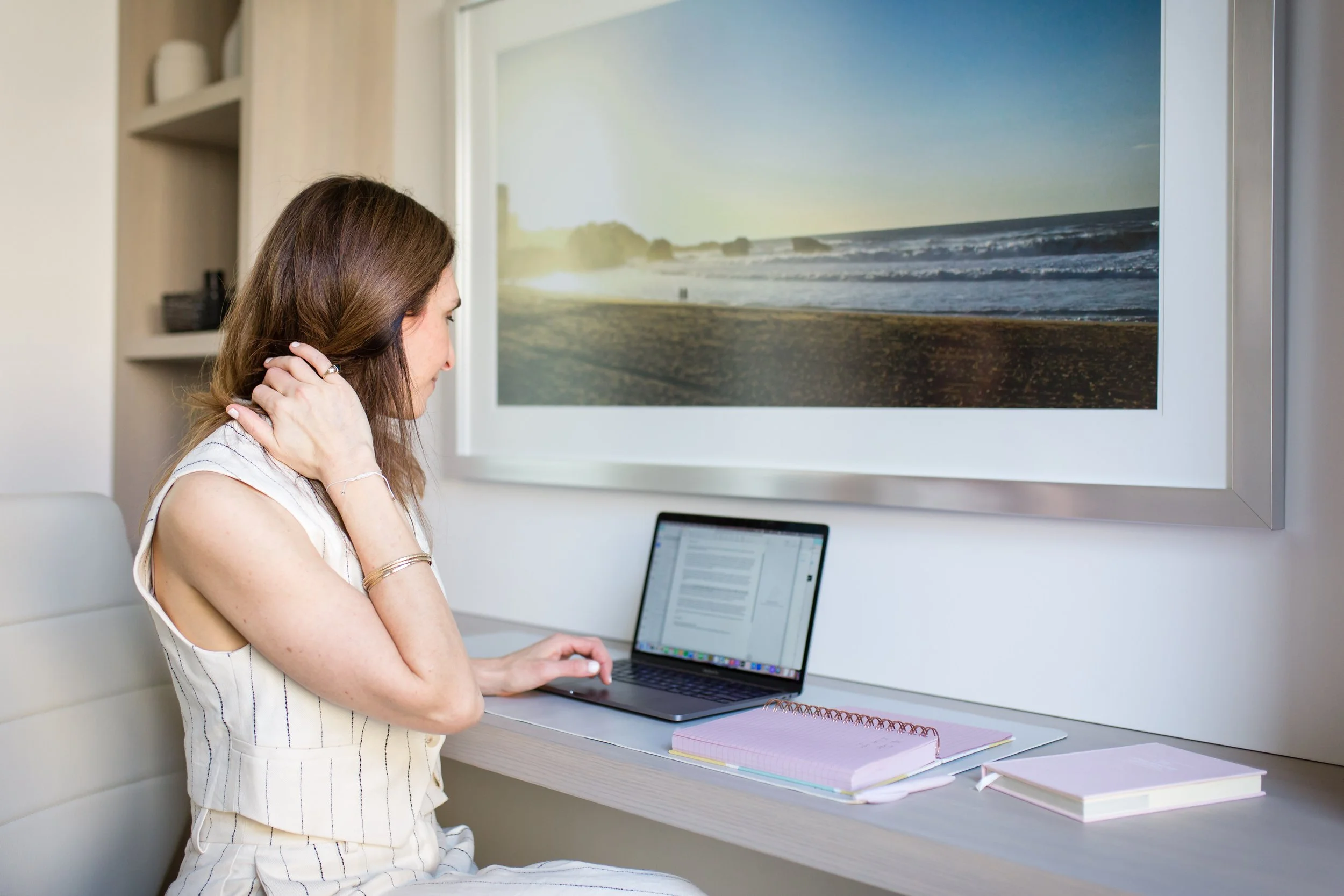 Woman sitting at a desk with a laptop, notebooks, and a large framed photo of a beach on the wall.