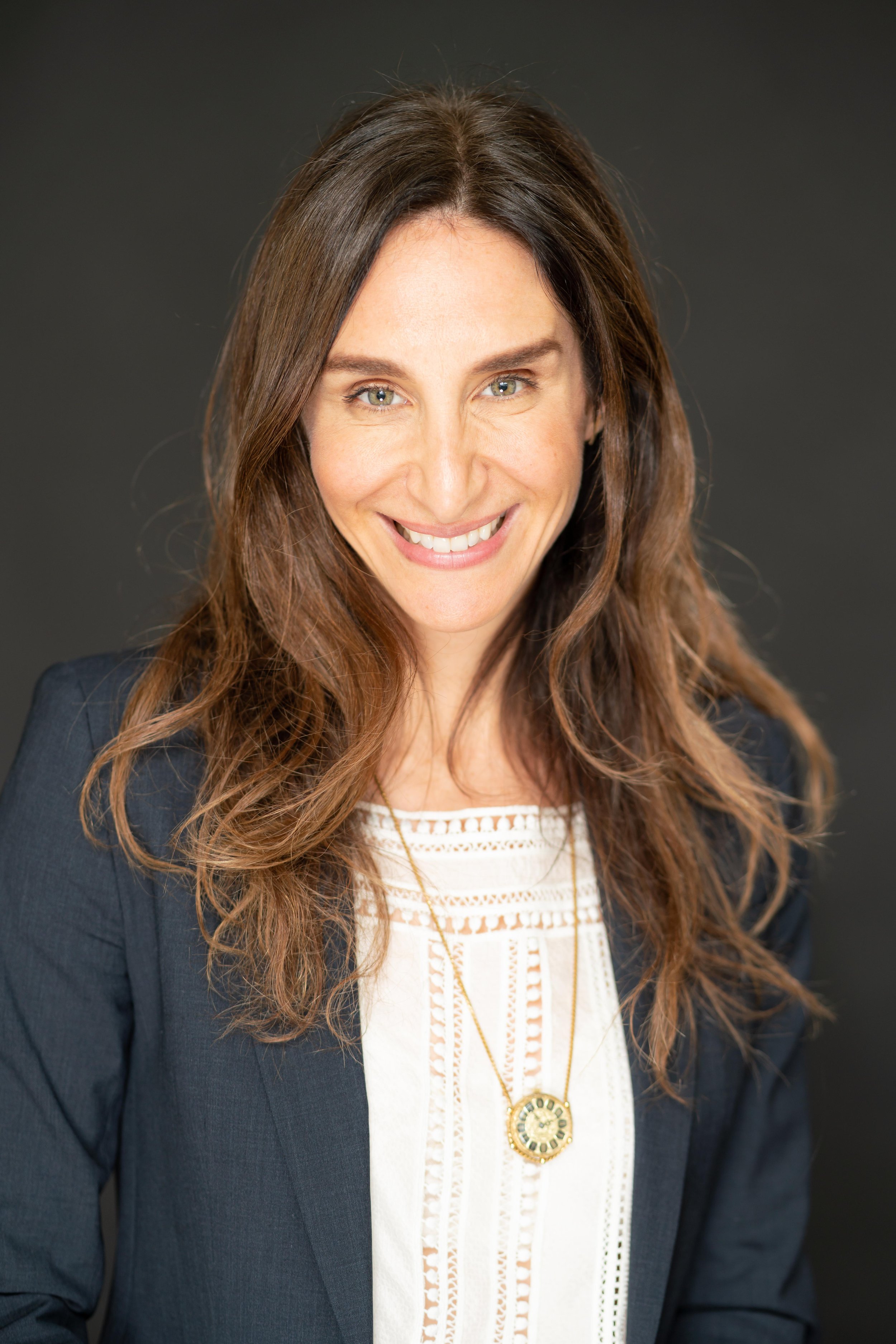 A woman with long brown hair and green eyes smiling, wearing a white top, a dark blazer, and a gold necklace, posed against a dark background.