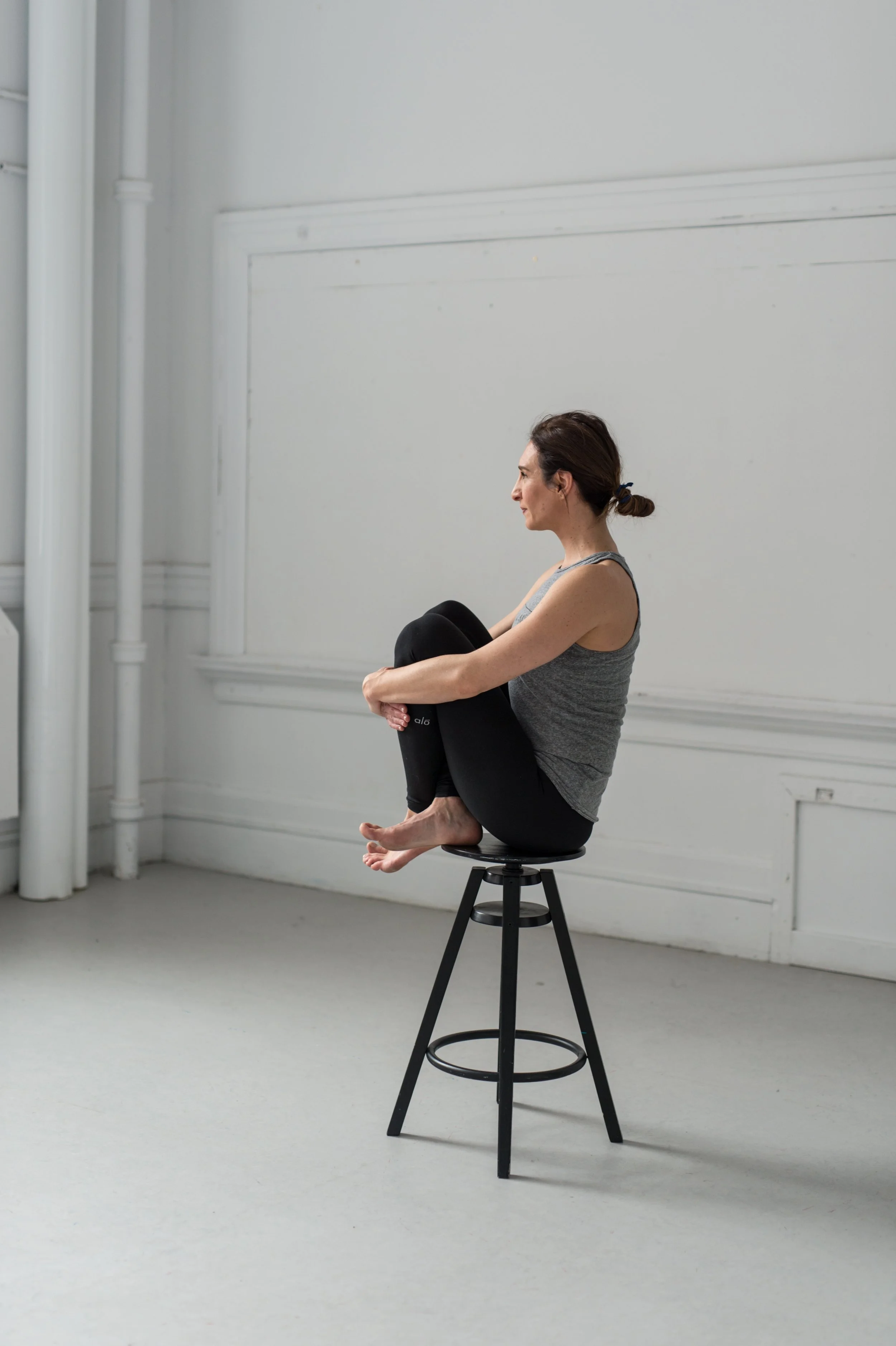 A woman sitting on a tall black stool inside a minimalist room, with her knees pulled to her chest and her arms wrapped around her legs, facing sideways.