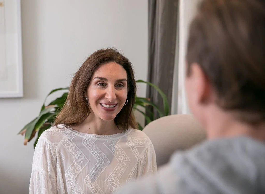 A woman with brown hair smiling and talking to a person with grayish hair in an indoor setting.