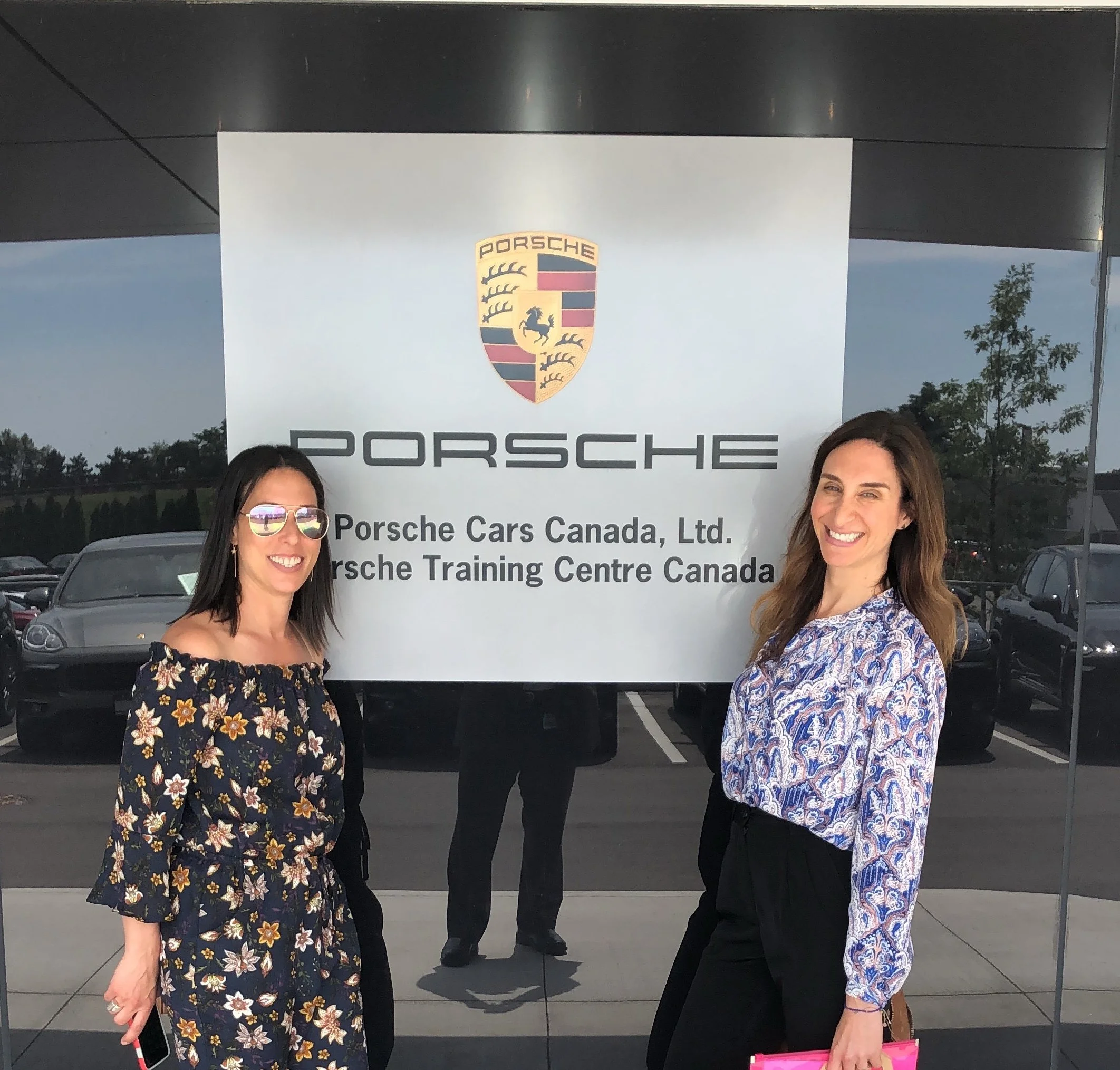 Two women smiling and standing in front of a Porsche sign at the Porsche Cars Canada Ltd. Porsche Training Centre Canada building. One woman is wearing sunglasses and a floral dress, and the other is wearing a patterned blouse and black pants.
