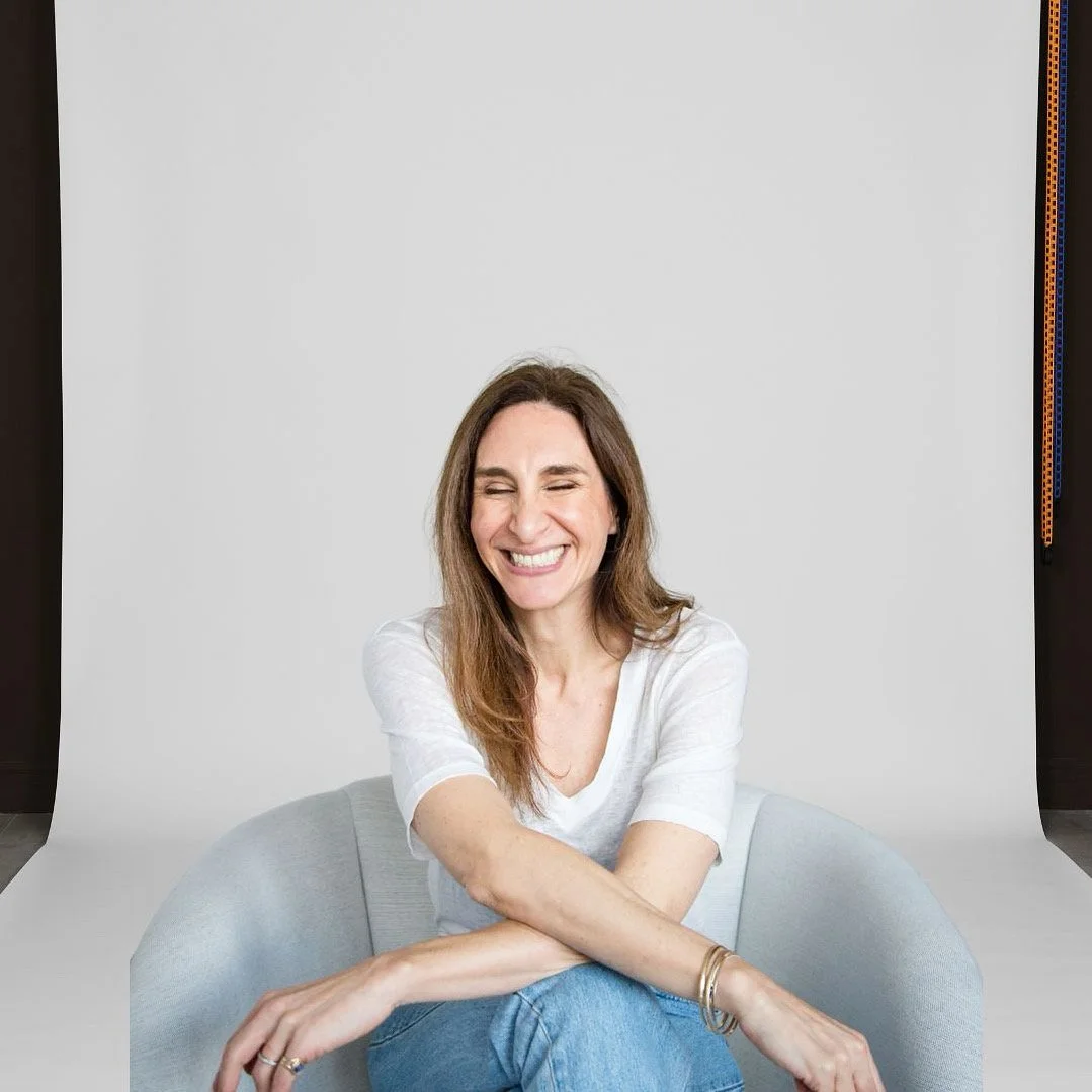 Sari Fox smiling in a white top, seated in a light grey chair