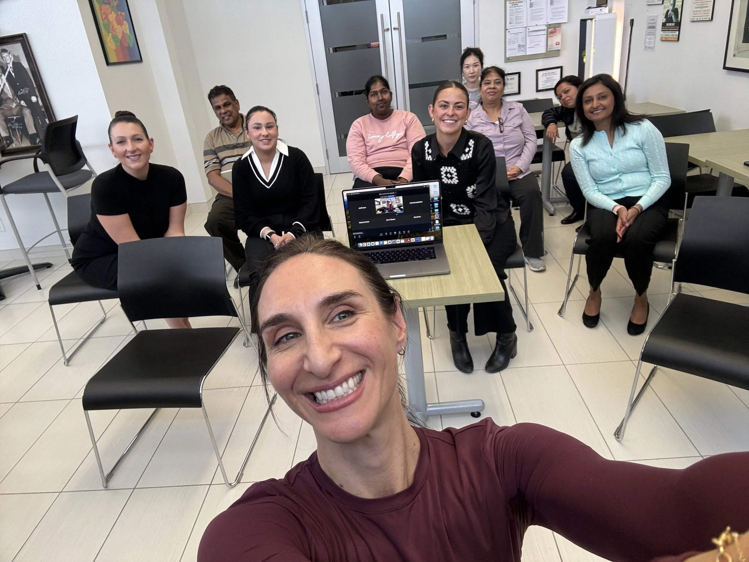 A woman taking a selfie with a group of nine diverse people seated behind her in a conference room. The group is smiling at the camera, with a laptop open on a small table in front of them. The room has framed certificates and pictures on the walls.