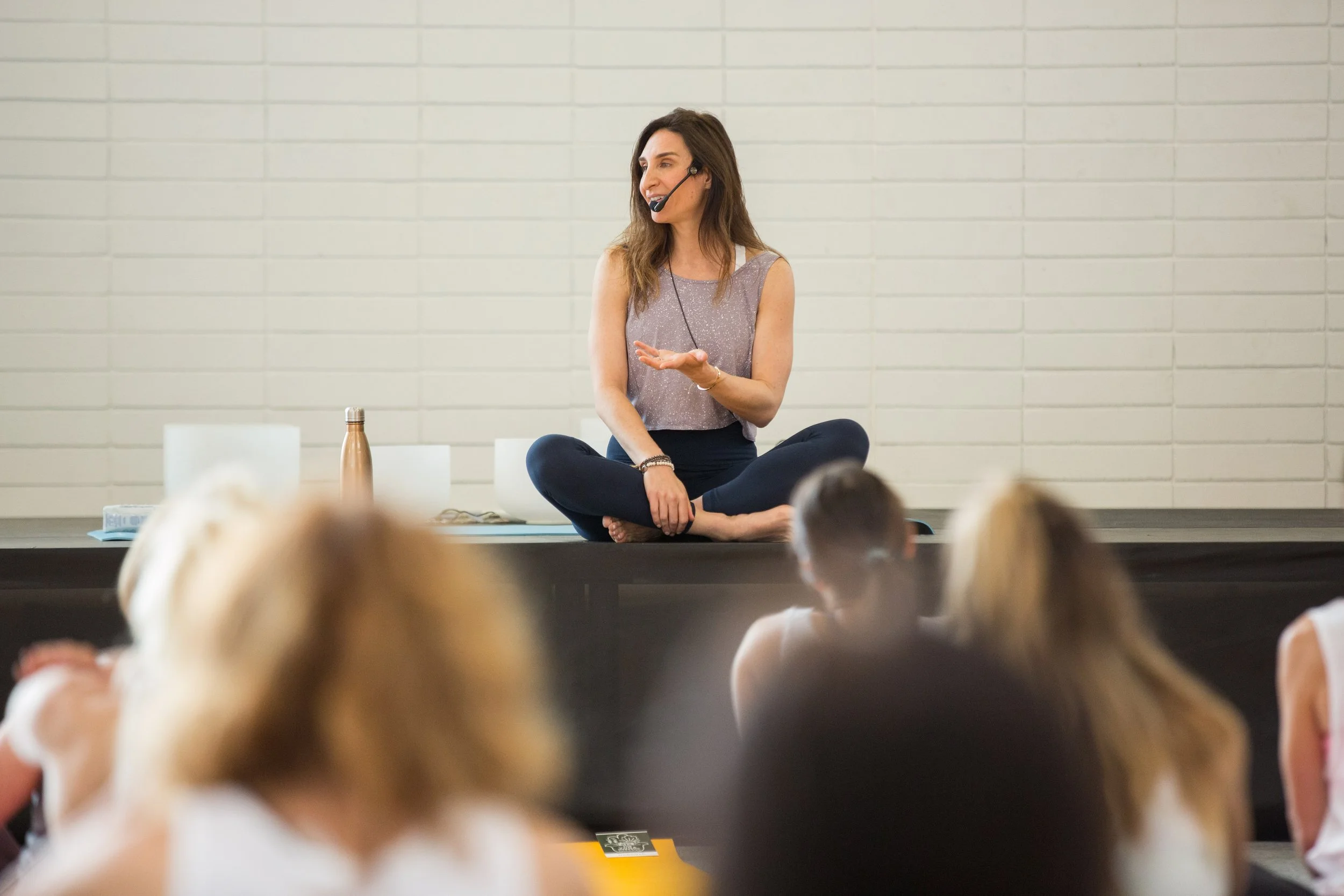 Woman sitting cross-legged on a table, speaking into a headset microphone, leading a workshop or class, with a group of people seated in front of her.