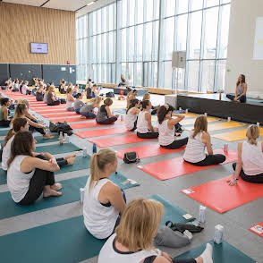 Group of women participating in a yoga class in a bright studio with large windows, seated on mats and following an instructor.