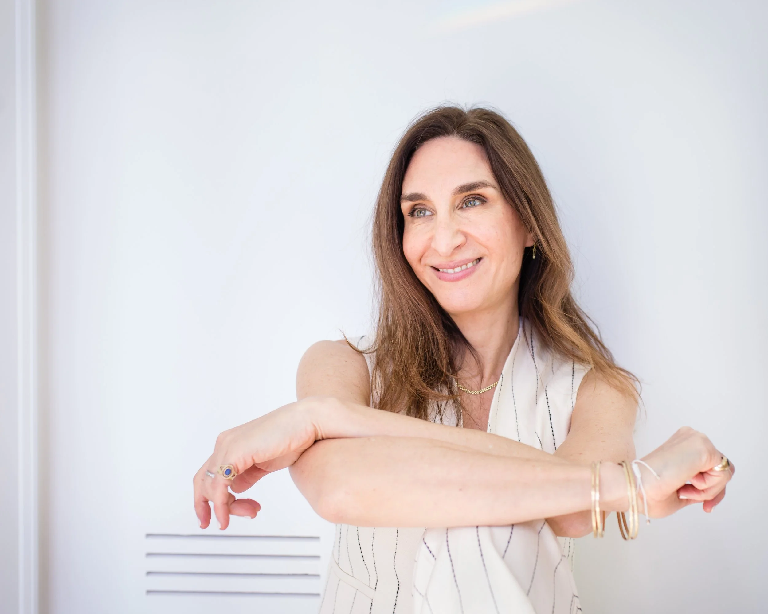A woman with long brown hair smiling while standing against a white wall, with her arms crossed and wearing jewelry.