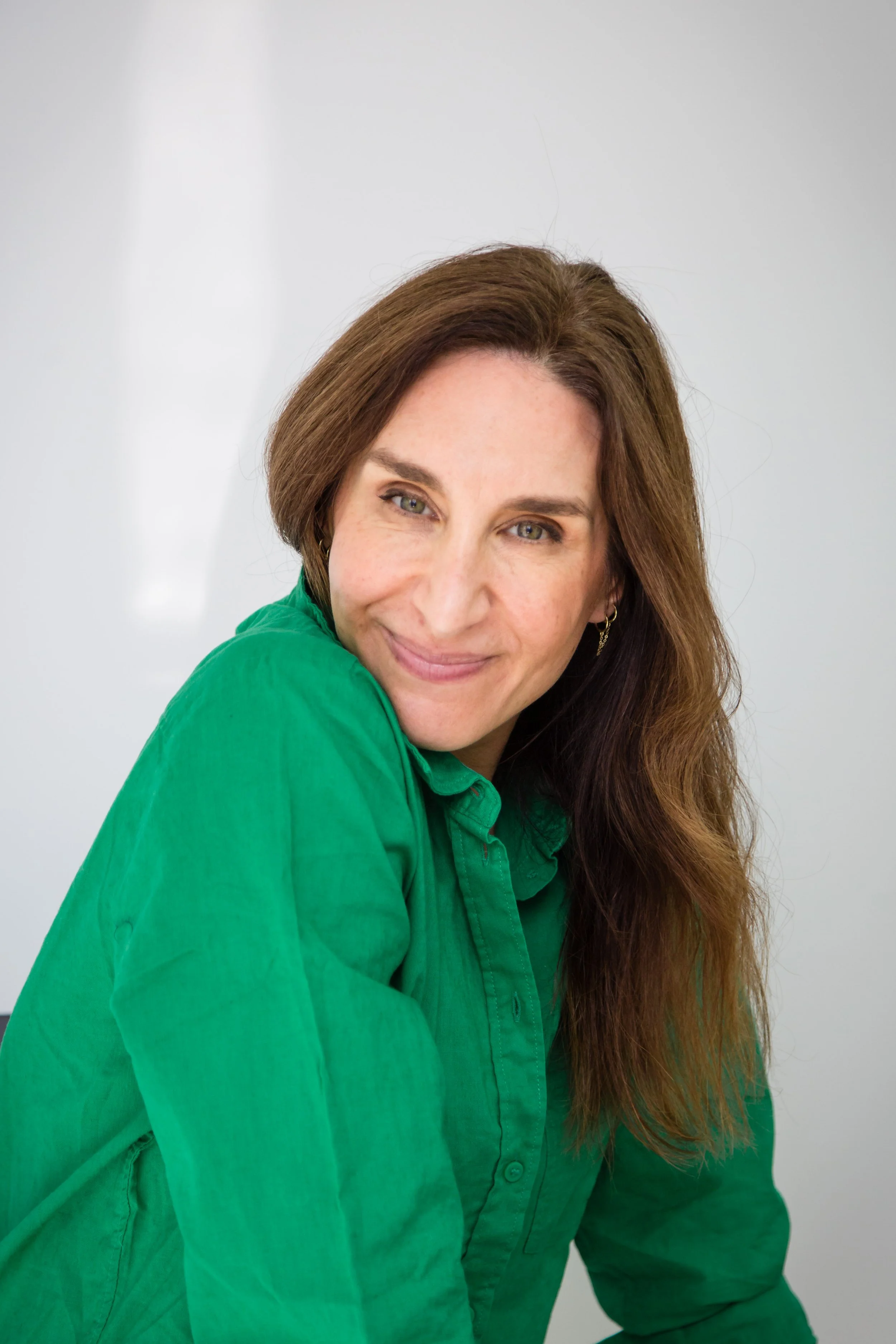 A woman with long brown hair wearing a green shirt, smiling softly at the camera against a plain background.