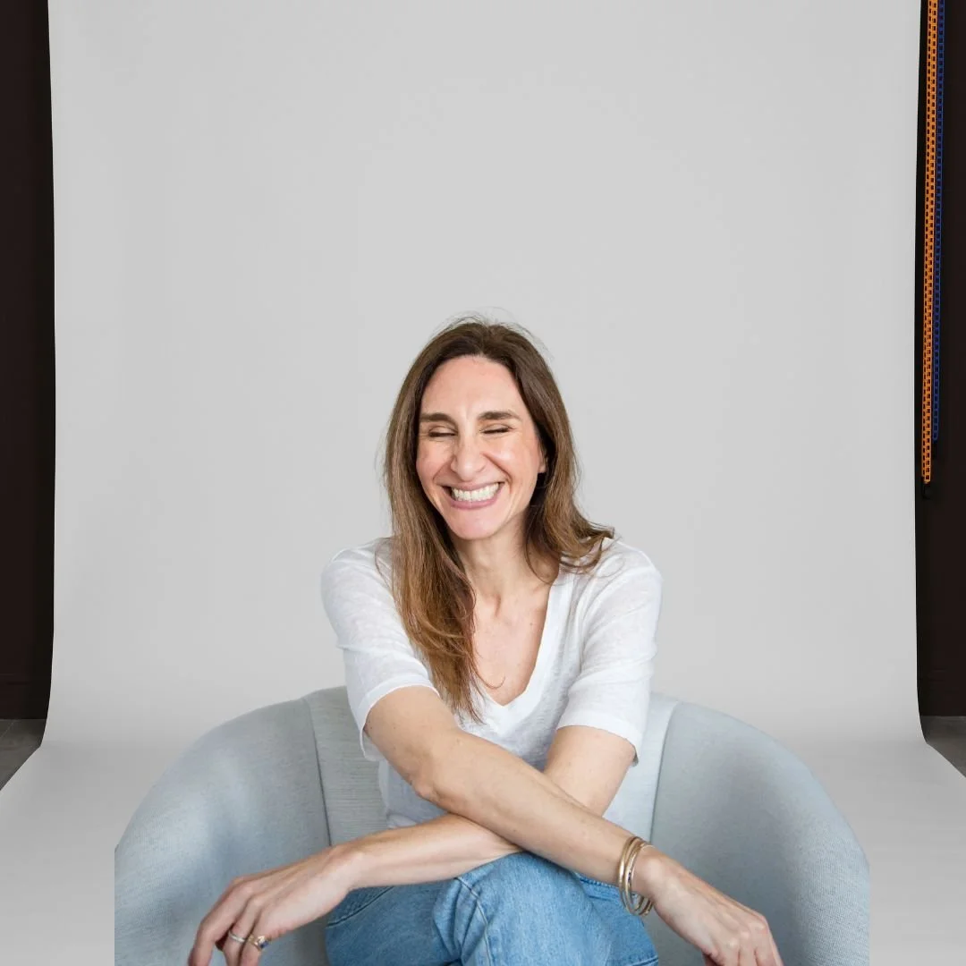 A woman with long brown hair, wearing a white T-shirt and blue jeans, laughing while sitting on a gray sofa in front of a plain gray backdrop.