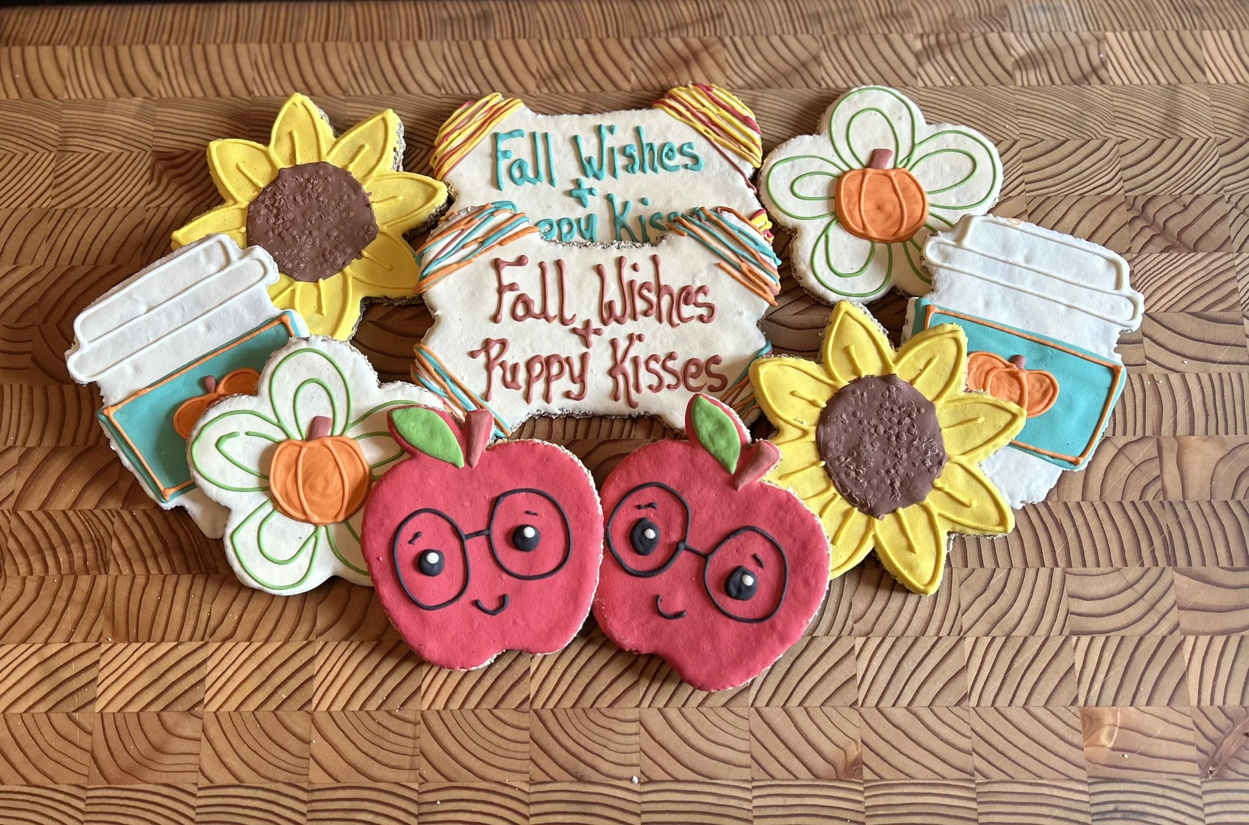 Assorted decorated fall-themed dog biscuits and cookies featuring sunflowers, pumpkins, apples, and cheerful messages from Day of the Dog Bakery, arranged on a wooden surface.
