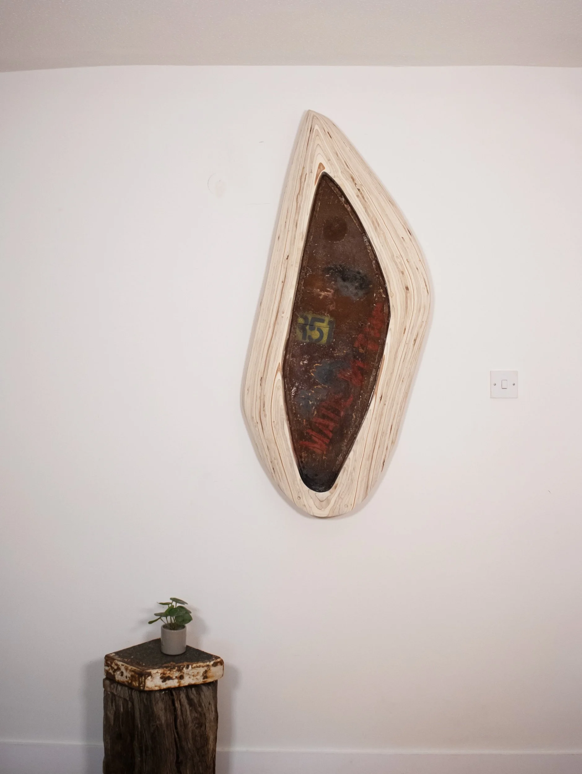 A minimalist interior wall decor featuring a wooden, boat-shaped shelf with a dark, rusty metal panel inside, mounted above a small plant on a rustic wooden block, with a white wall and a light switch in the background.