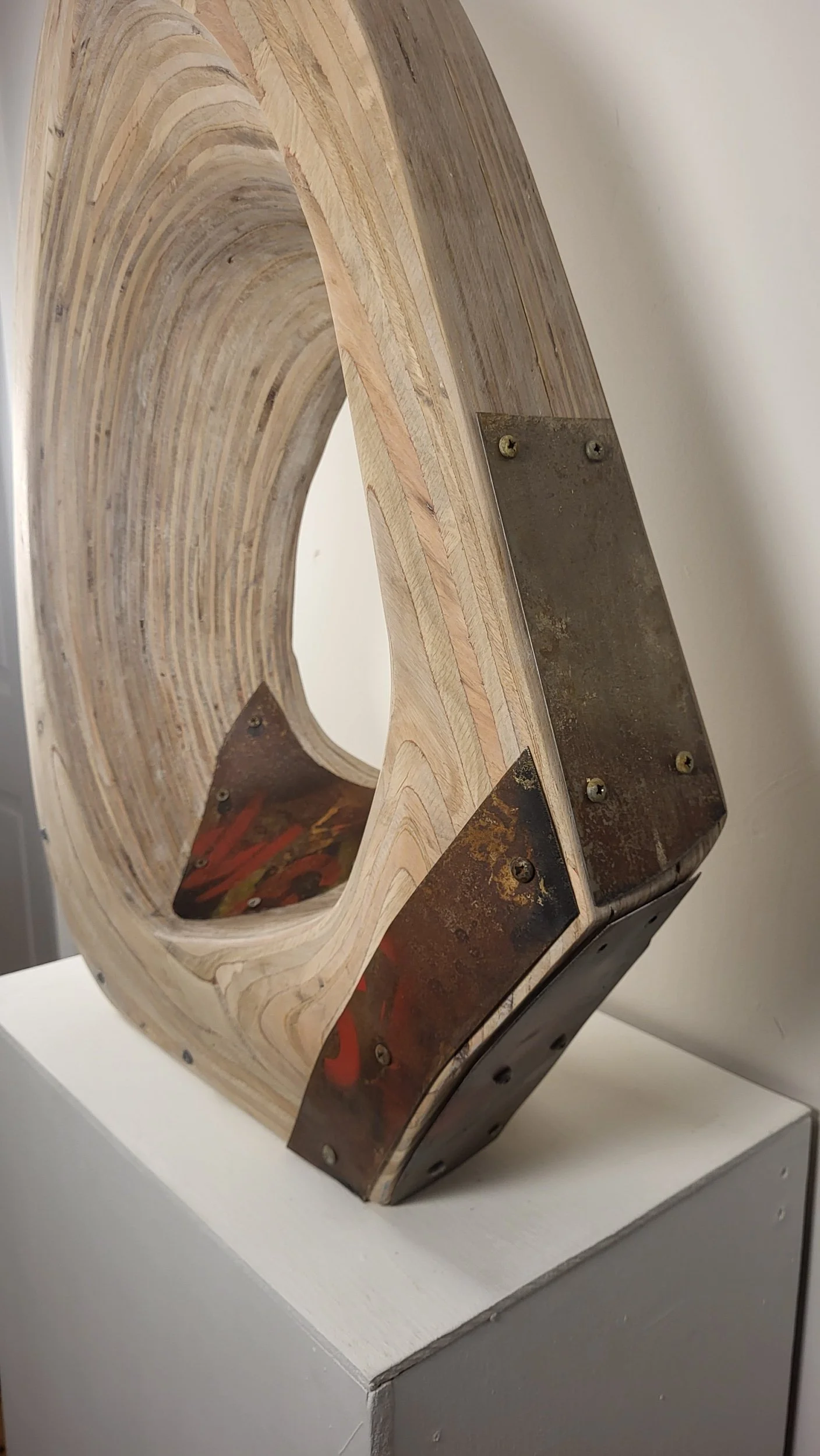 A sculpture made of layered wood with a curved shape, mounted on a white pedestal. The sculpture has metal plates and screws attached to it, with visible rust and red paint marks.