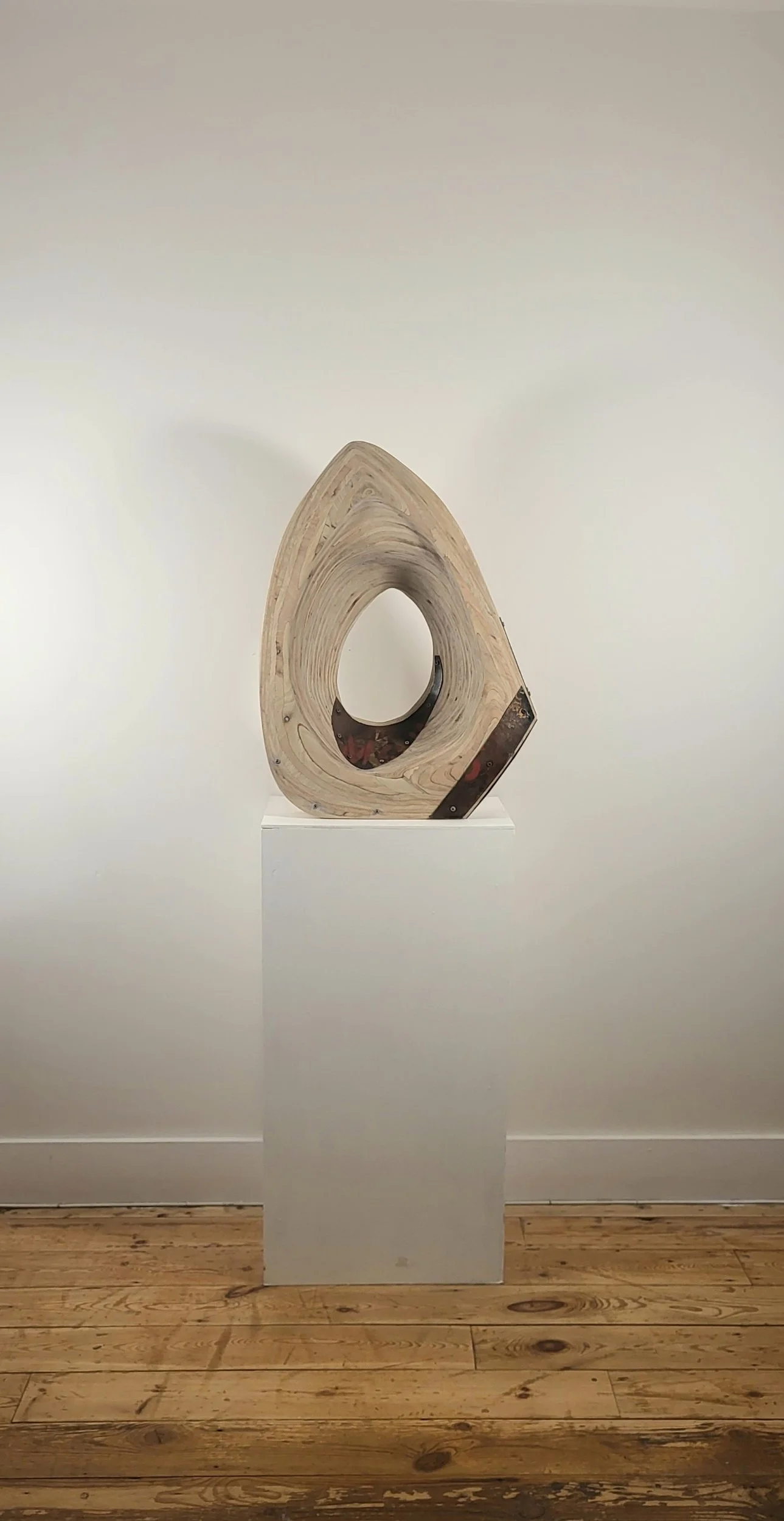 A modern wooden sculpture with a central hole, displayed on a white pedestal against a plain white wall.