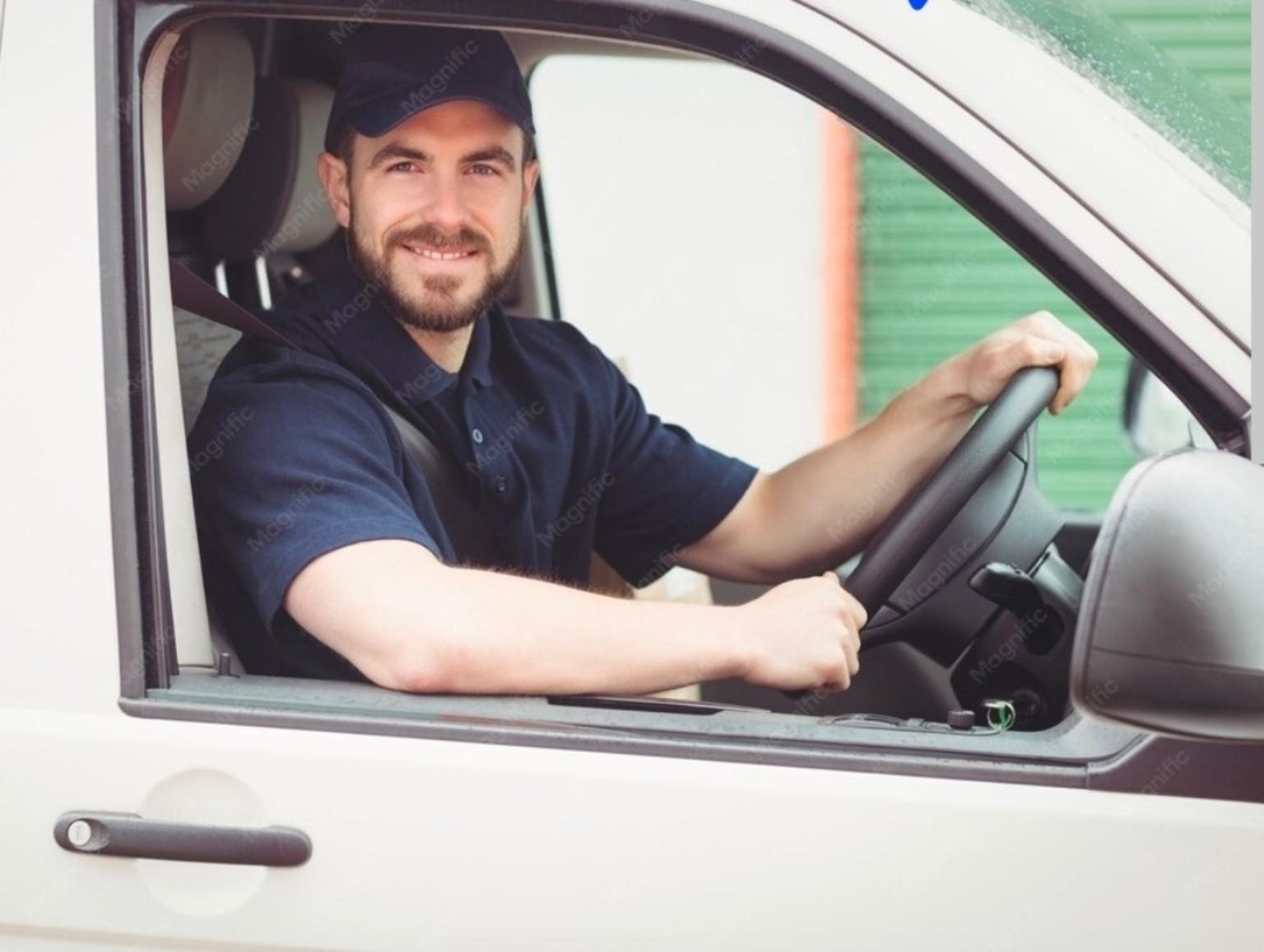 A man with a beard and wearing a navy cap and navy polo shirt is sitting in the driver's seat of a white vehicle, smiling and holding the steering wheel.