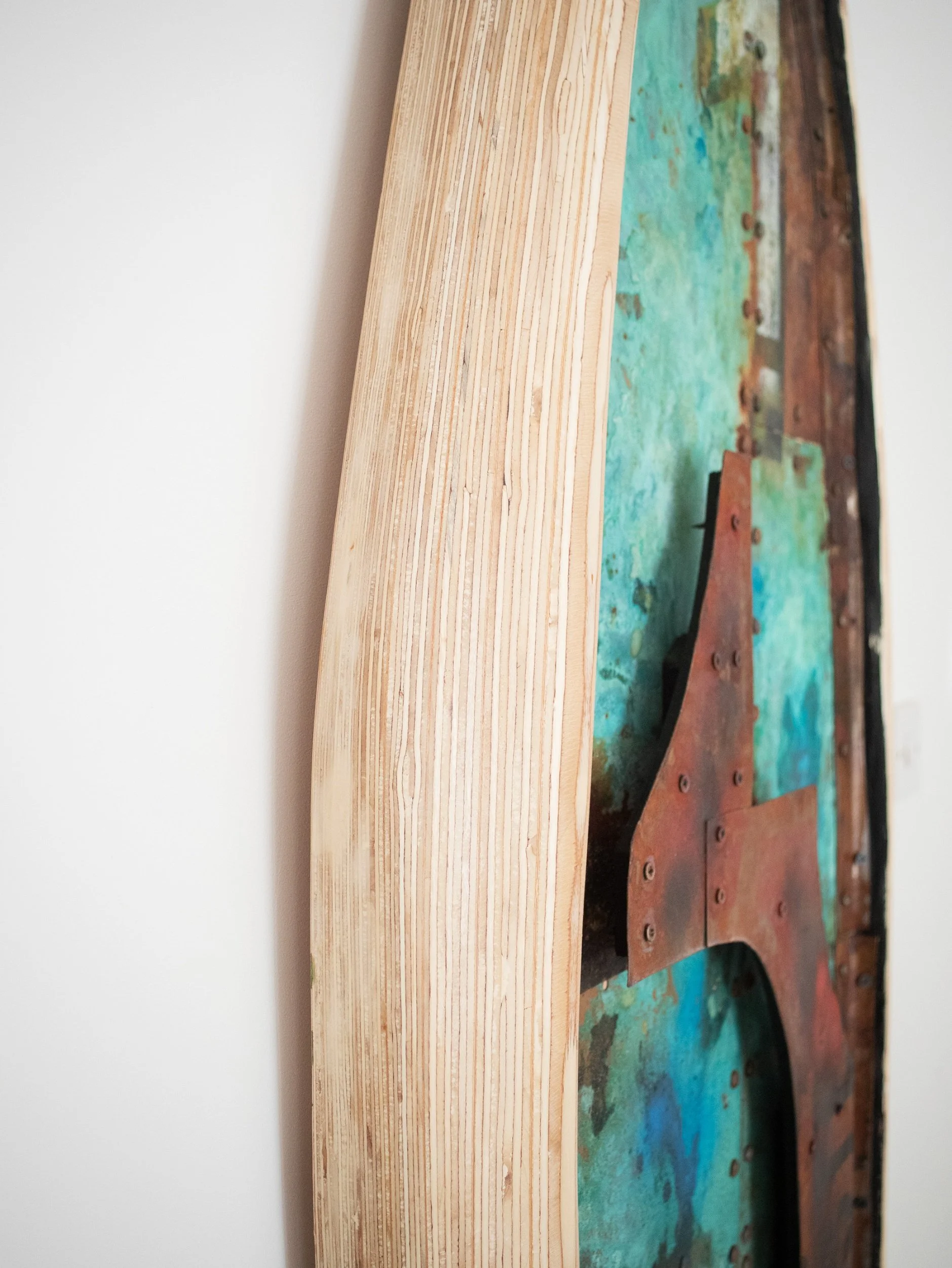 Close-up of an abstract painting with shades of green, blue, and brown, mounted on a wooden frame, leaning against a white wall.