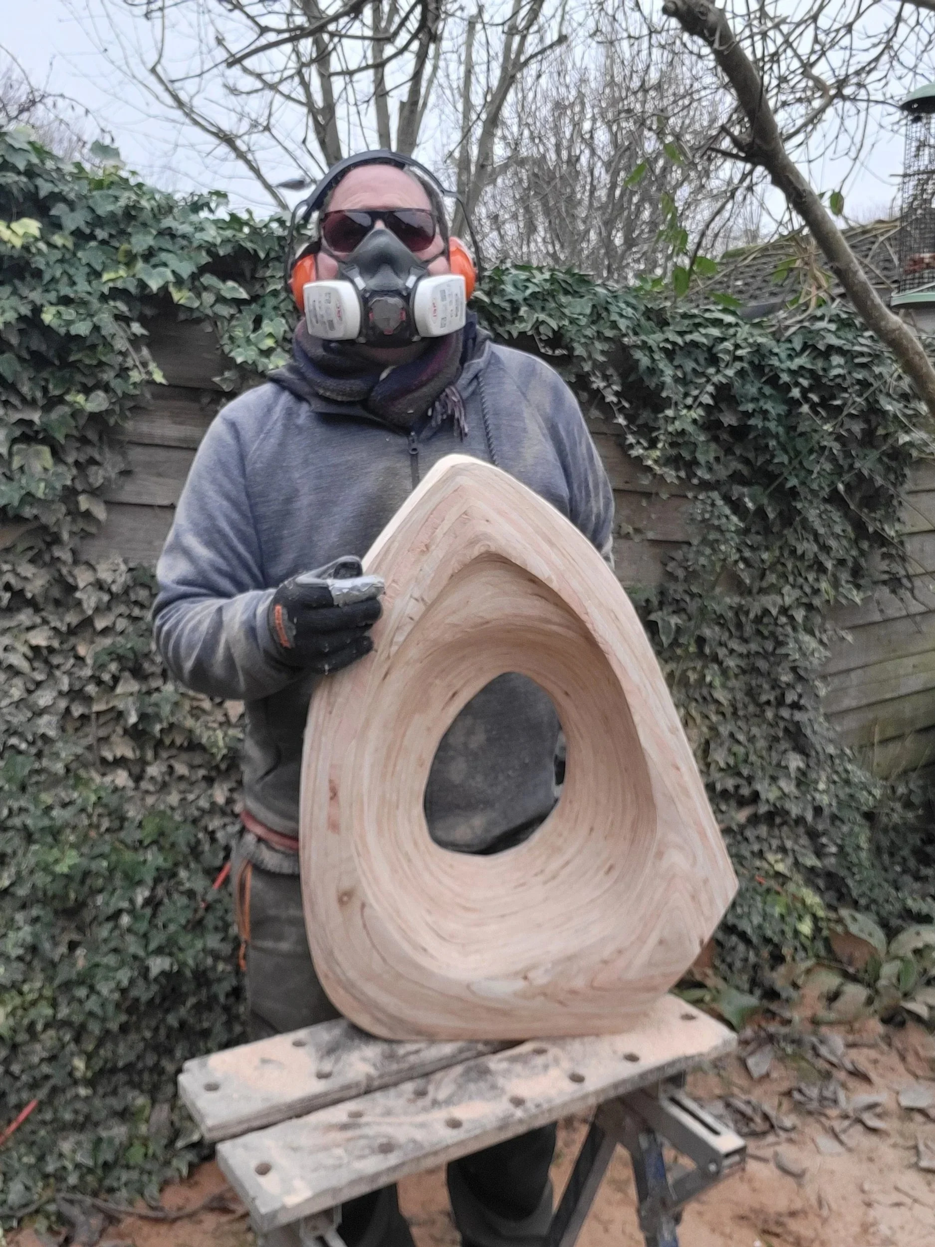 A person wearing safety goggles, ear protection, a dust mask, and gloves holding a large wooden ring-shaped sculpture outdoors. The sculpture is placed on a workbench in a garden with leafless trees and a wooden fence in the background.