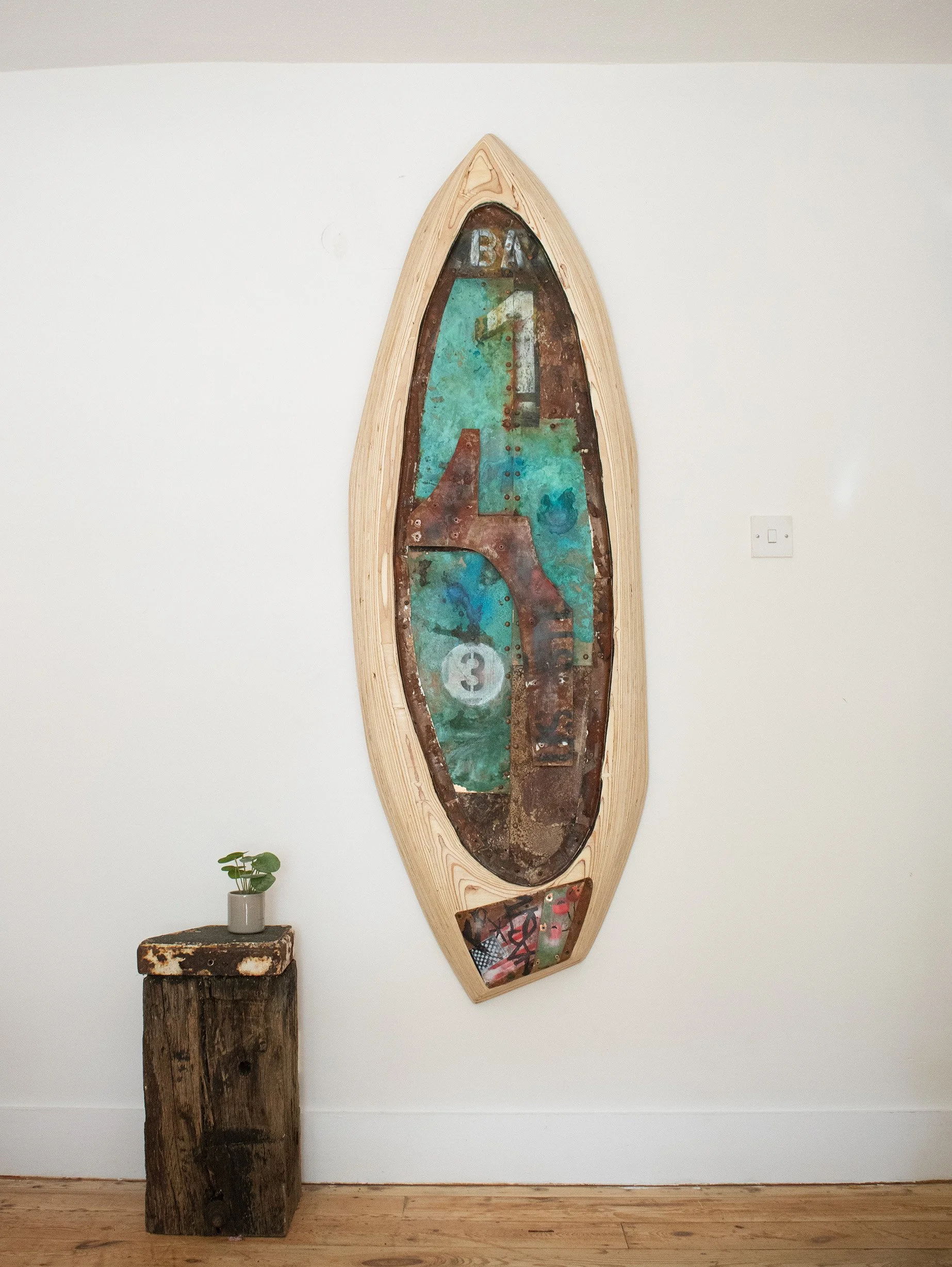 A mixed media art piece mounted on a wall, shaped like an elongated oval or surfboard, featuring a rusty, weathered metal surface with painted numbers and abstract designs. A small potted plant is placed on a rustic wooden pedestal below it.